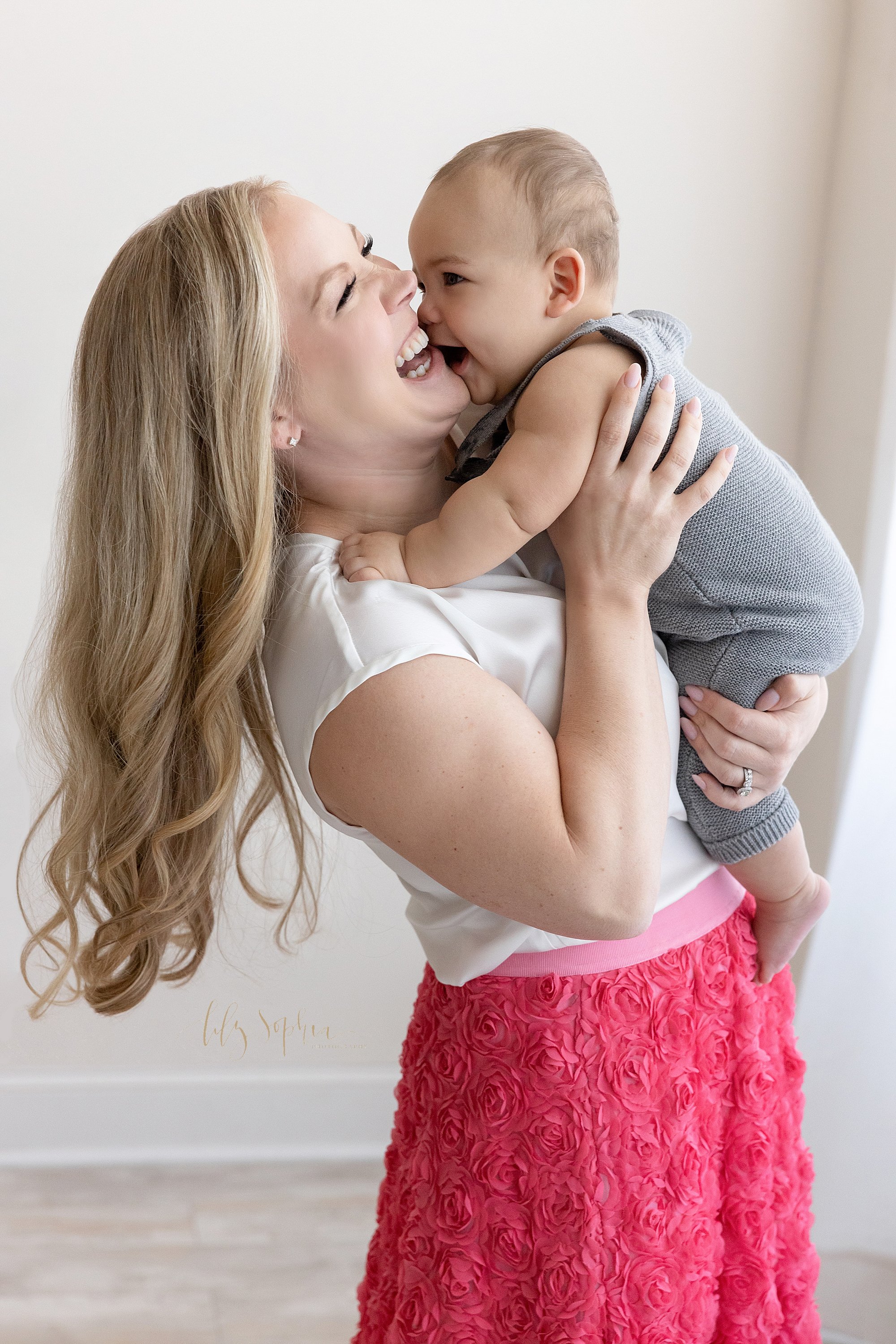  Baby photo of a mother holding her baby boy as he tries to kiss her taken next to a window streaming natural light near Sandy Springs in Atlanta in a photography studio. 