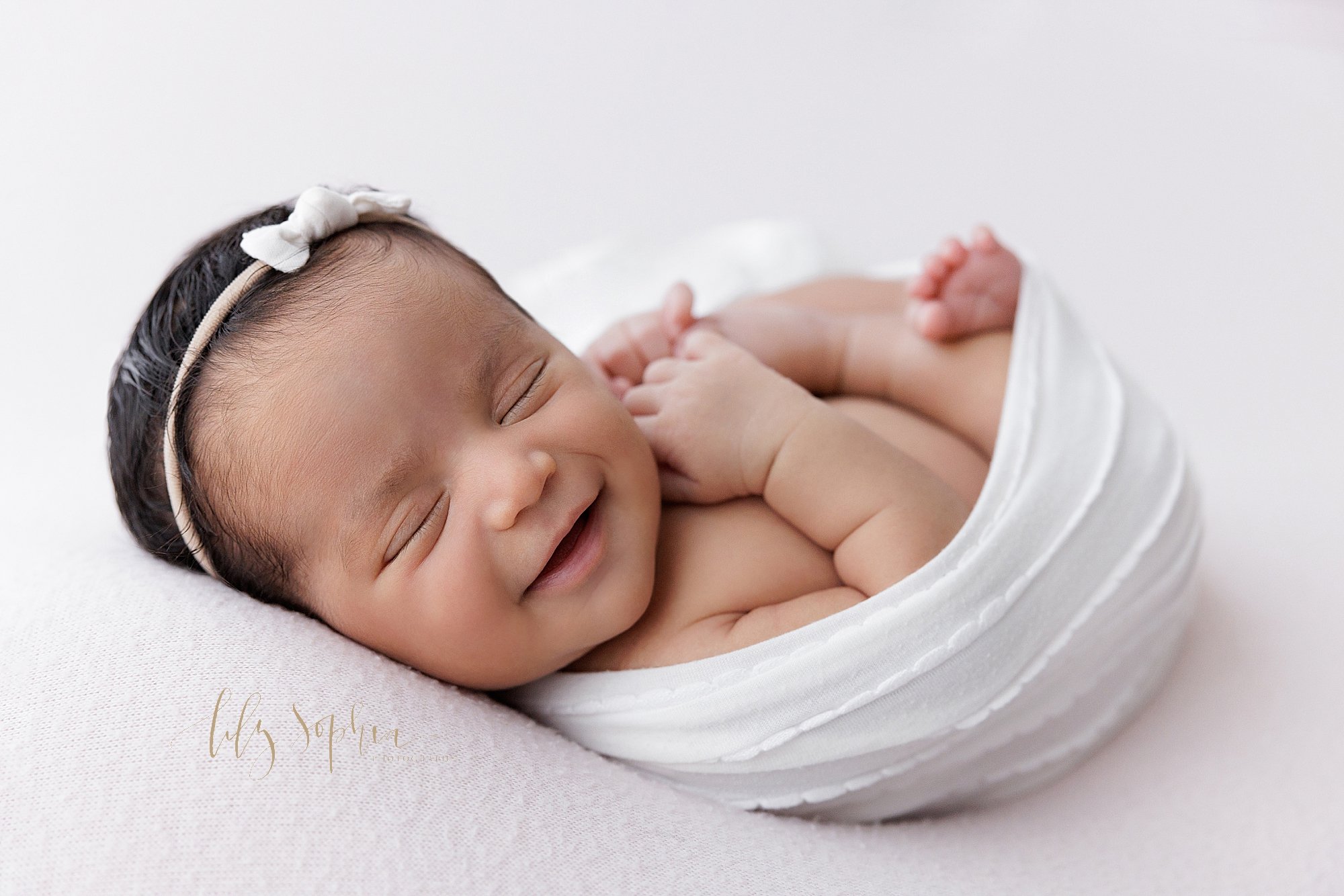  Newborn portrait of a smiling newborn baby girl wearing a bow headband in her hair while she is tucked into a stretchy swaddle taken near Cumming in Atlanta in a natural light studio. 