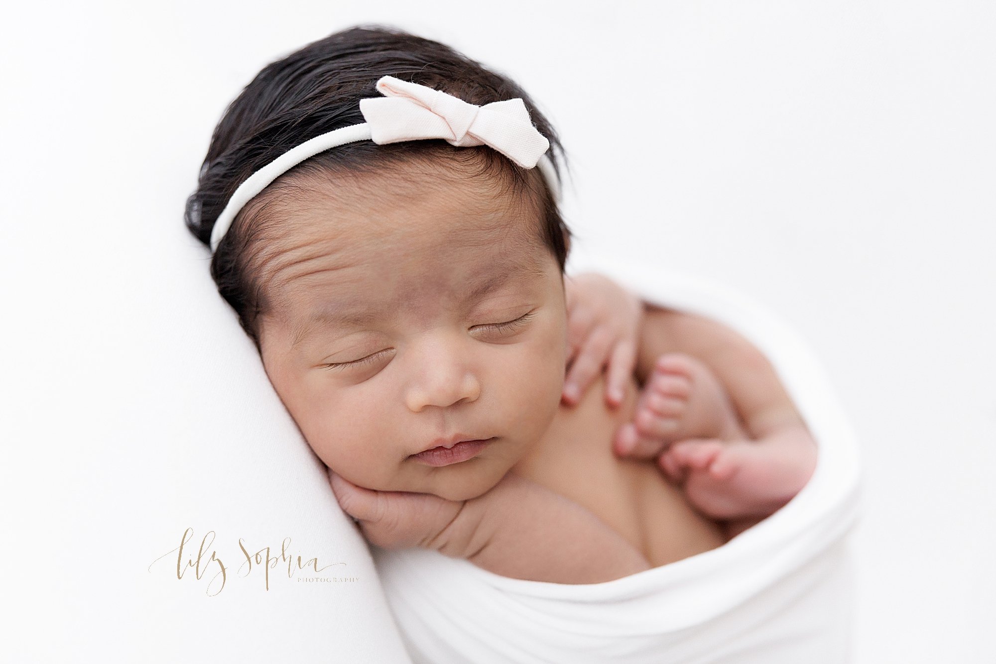  Newborn portrait of a newborn Indian girl wearing a bow headband in her jet black hair as she dreams and raises her eyebrows while cradled in a stretchy swaddle taken in natural light near Roswell in Atlanta in a photography studio. 