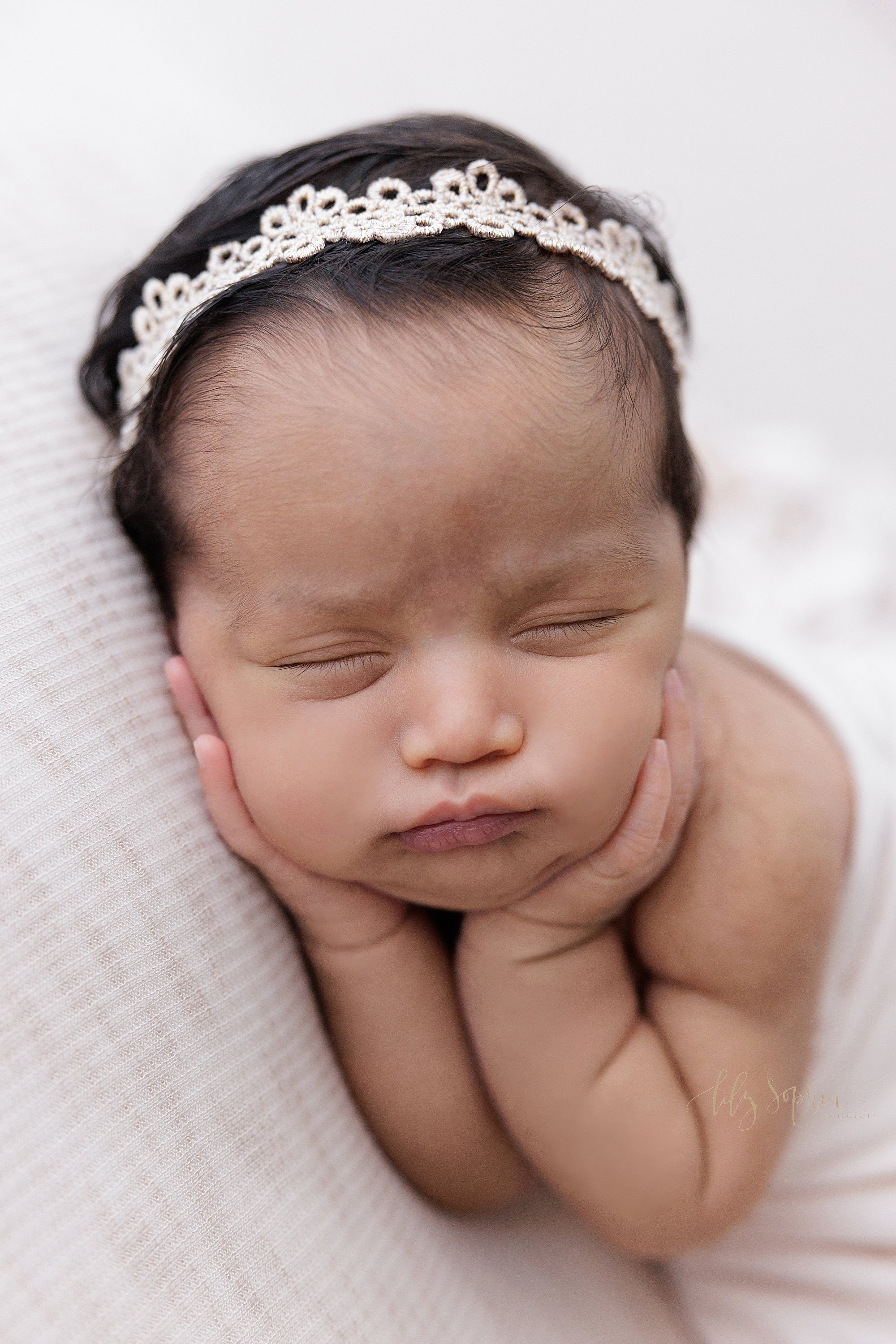  Newborn picture of a newborn Indian baby girl wearing a delicate stretchy headband in her hair with her hands holing her cheeks as she pouts in her sleep while turning her head to her right taken using natural light near Vinings in Atlanta in a phot