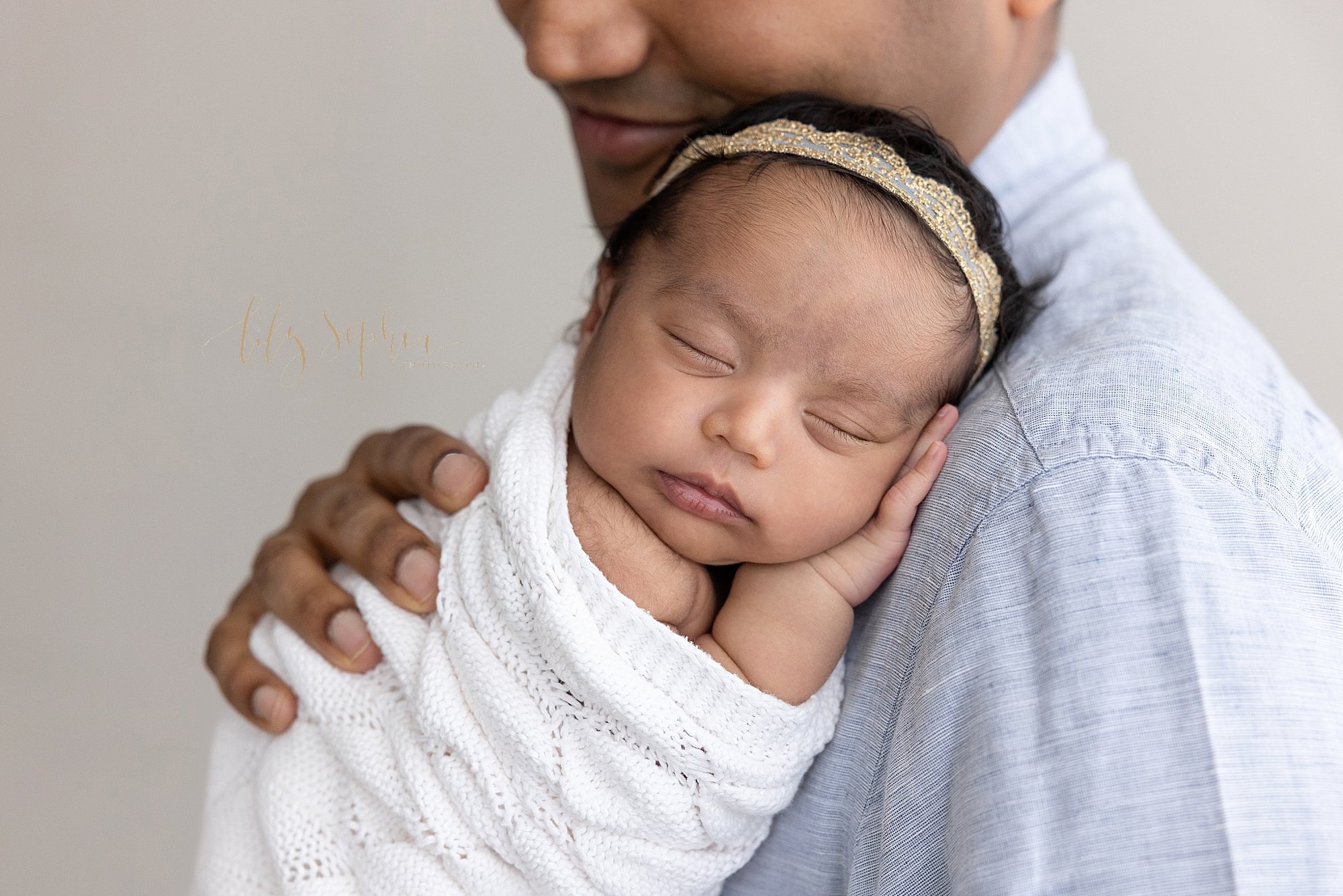  Close-up photo of a newborn baby girl wrapped in a white crocheted blanket as she sleeps against her father’s shoulder taken near Ansley Park in Atlanta in a studio using natural light. 