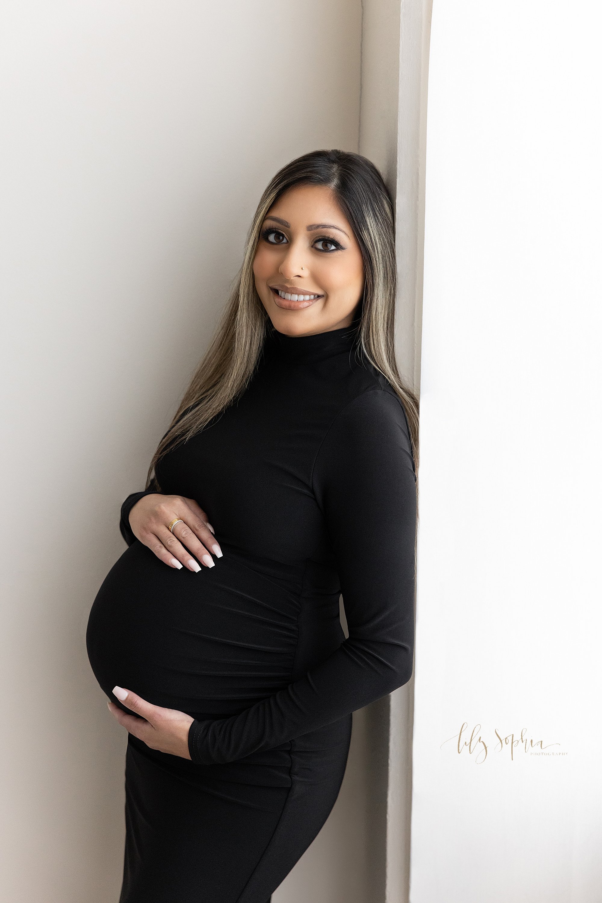  Maternity portrait of a happy expectant mother as she stands with her back against a wall next to a window streaming natural light and frames her belly with her hands taken in a photography studio near Brookhaven in Atlanta. 