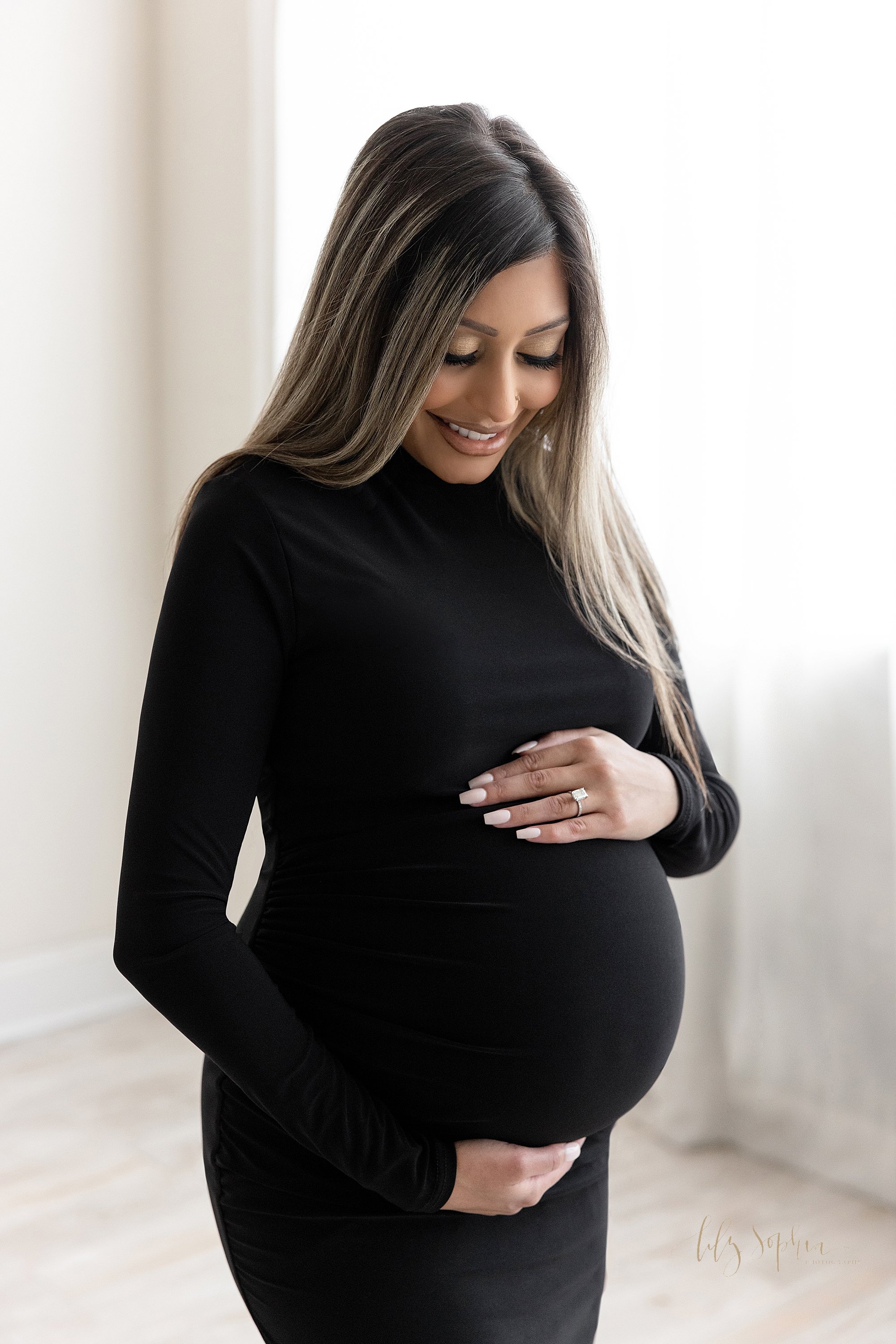  Maternity portrait of an expectant mother standing next to a window streaming natural light as she frames her belly with her hands and smiles at the baby kicking inside her taken near Decatur in Atlanta, Georgia. 