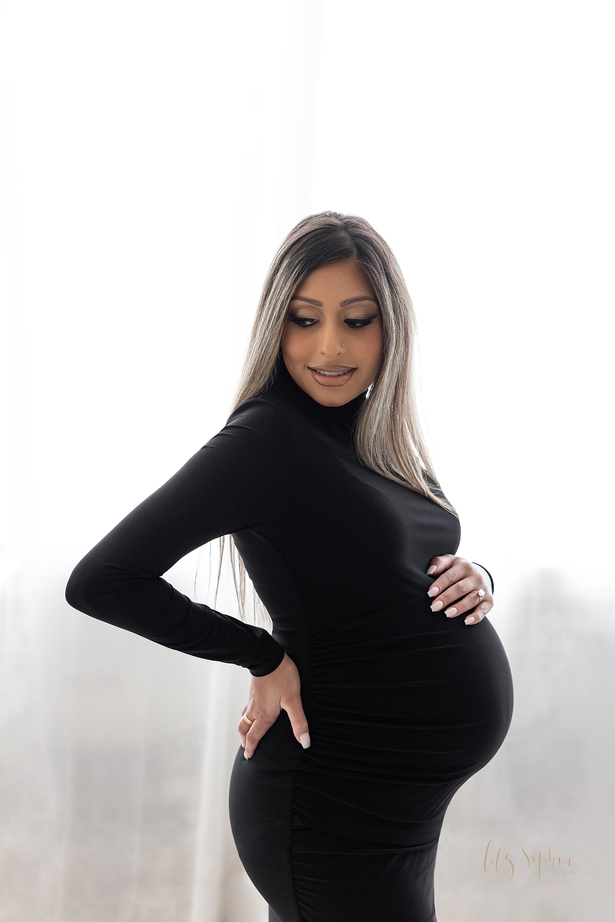  Maternity photo session with a pregnant mother standing in front of a window streaming natural light and wearing a knit long sleeve turtle neck gown with her right hand on her hip and her left hand on the top of her belly as she looks over her right
