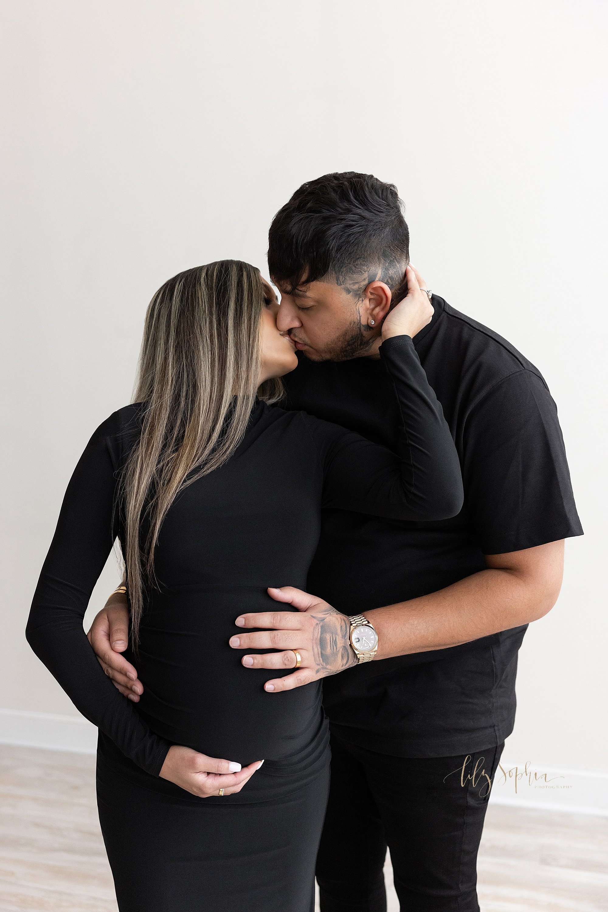  Maternity photo shoot of a pregnant mother standing with her right hand holding the base of her belly as her husband stands behind her and she grabs his neck with her left hand as the couple kiss while he places his right hand on their child in uter