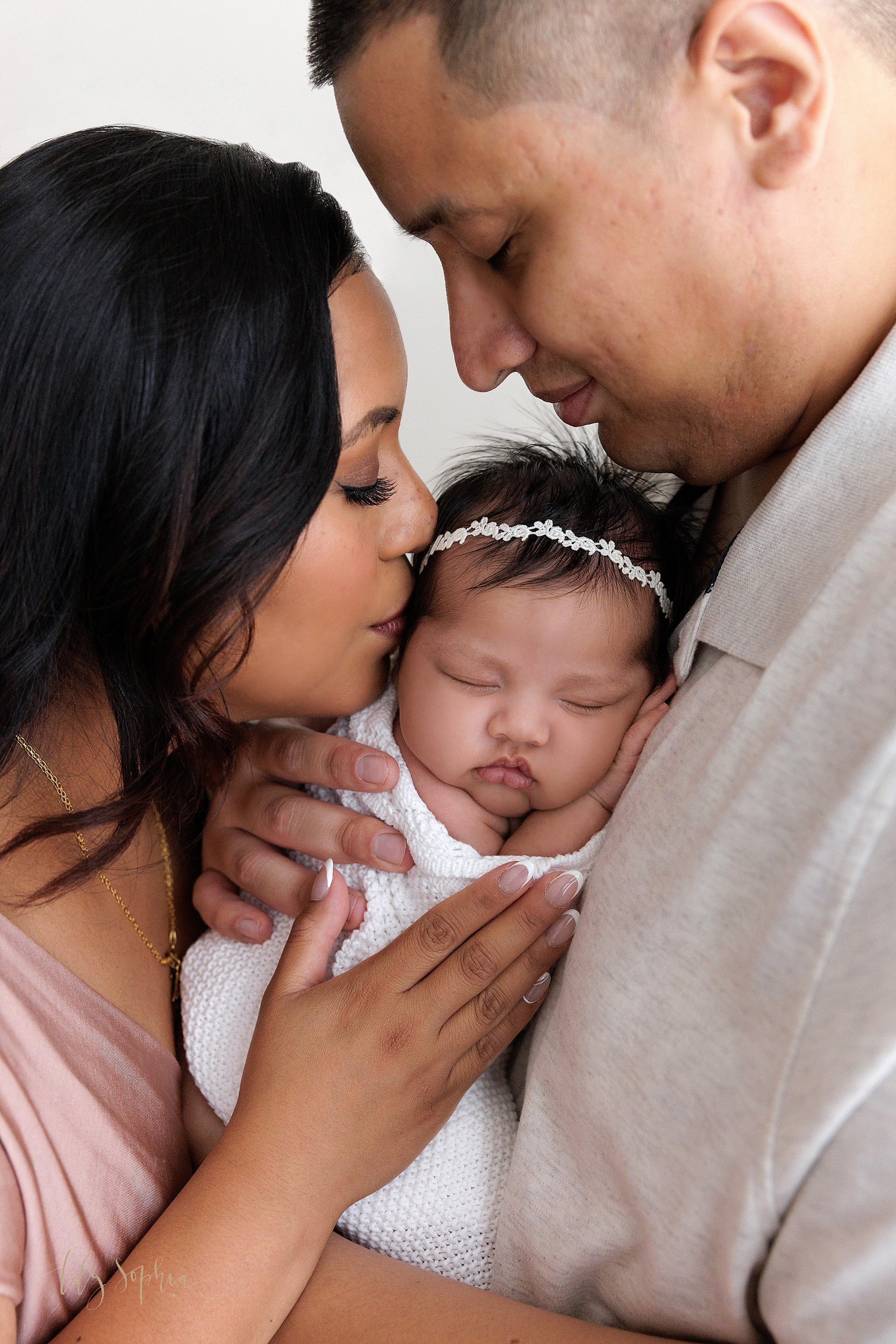  Close-up newborn portrait of a peacefully sleeping newborn baby girl being held against her father’s chest as her mother faces her father and kisses the side of her head taken in a natural light studio near Smyrna in Atlanta, Georgia.  