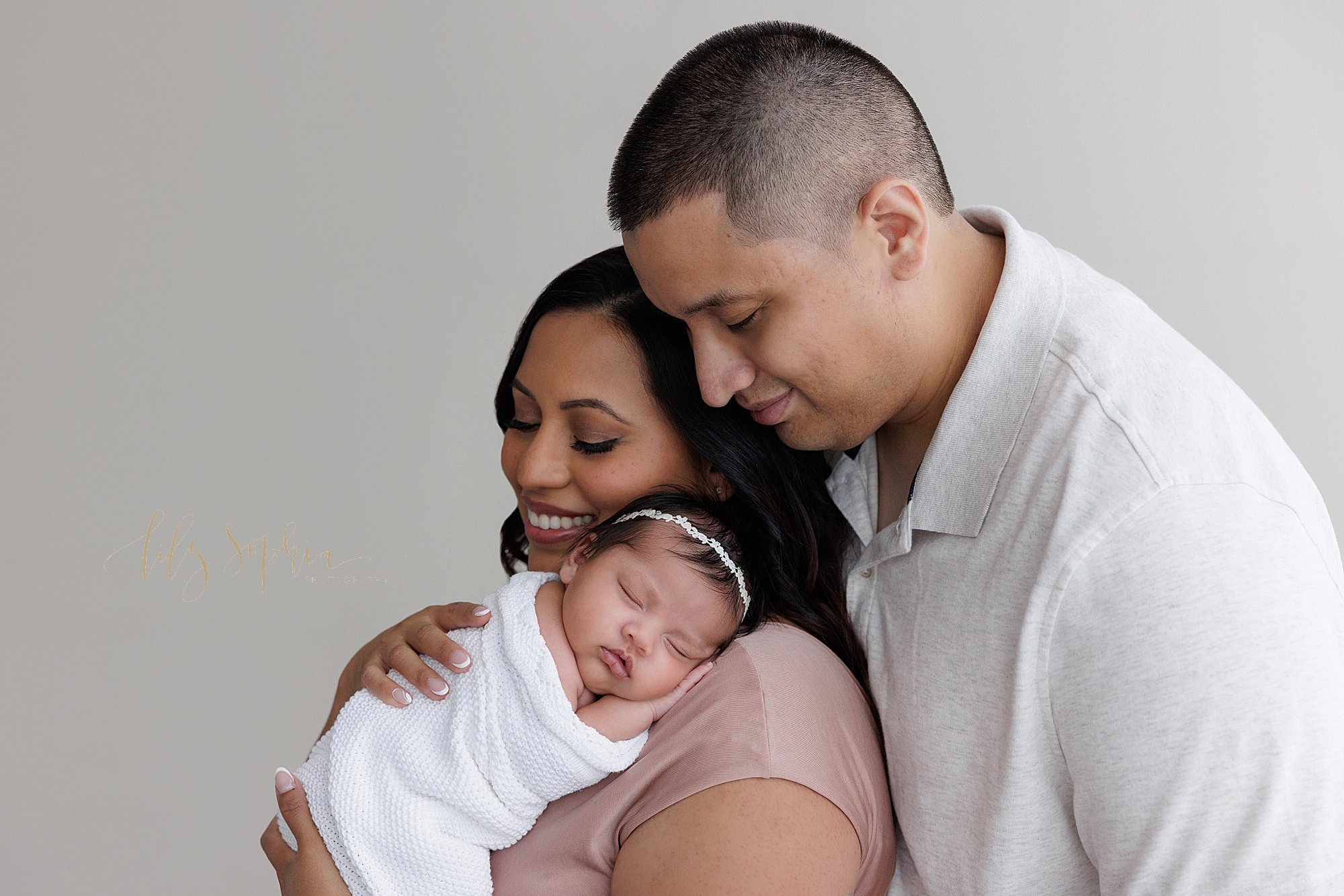  Newborn portrait of a mother holding her sleeping newborn baby girl on her shoulder as her husband stands behind her and looks on taken near Roswell in Atlanta in a studio that uses natural light. 