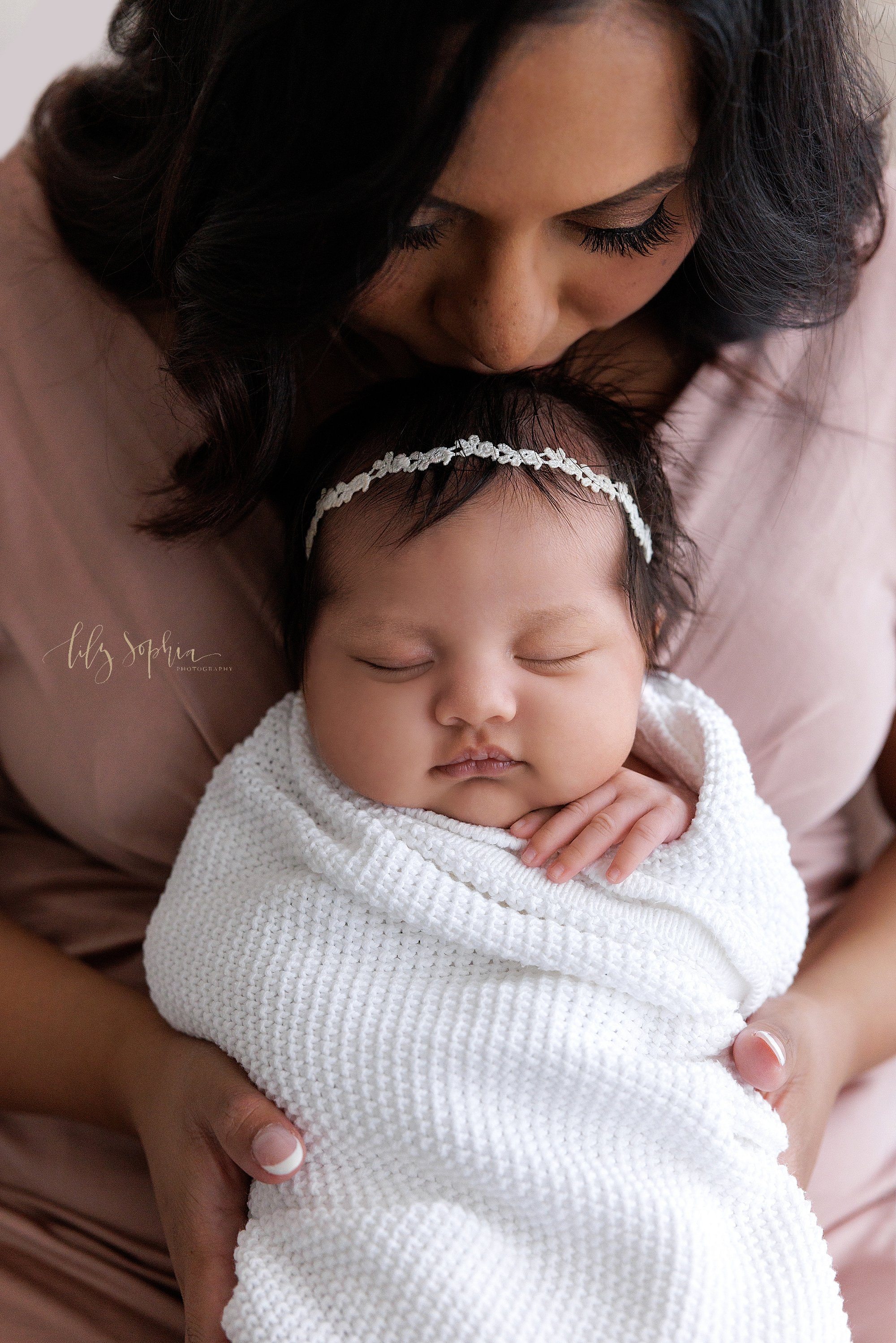  Newborn portrait or a newborn baby girl wrapped in a soft white blanket and wearing a delicate laurel headband in her black hair as her mother holds her on her lap and kisses the crown of her head taken in a studio that uses natural light near Morni