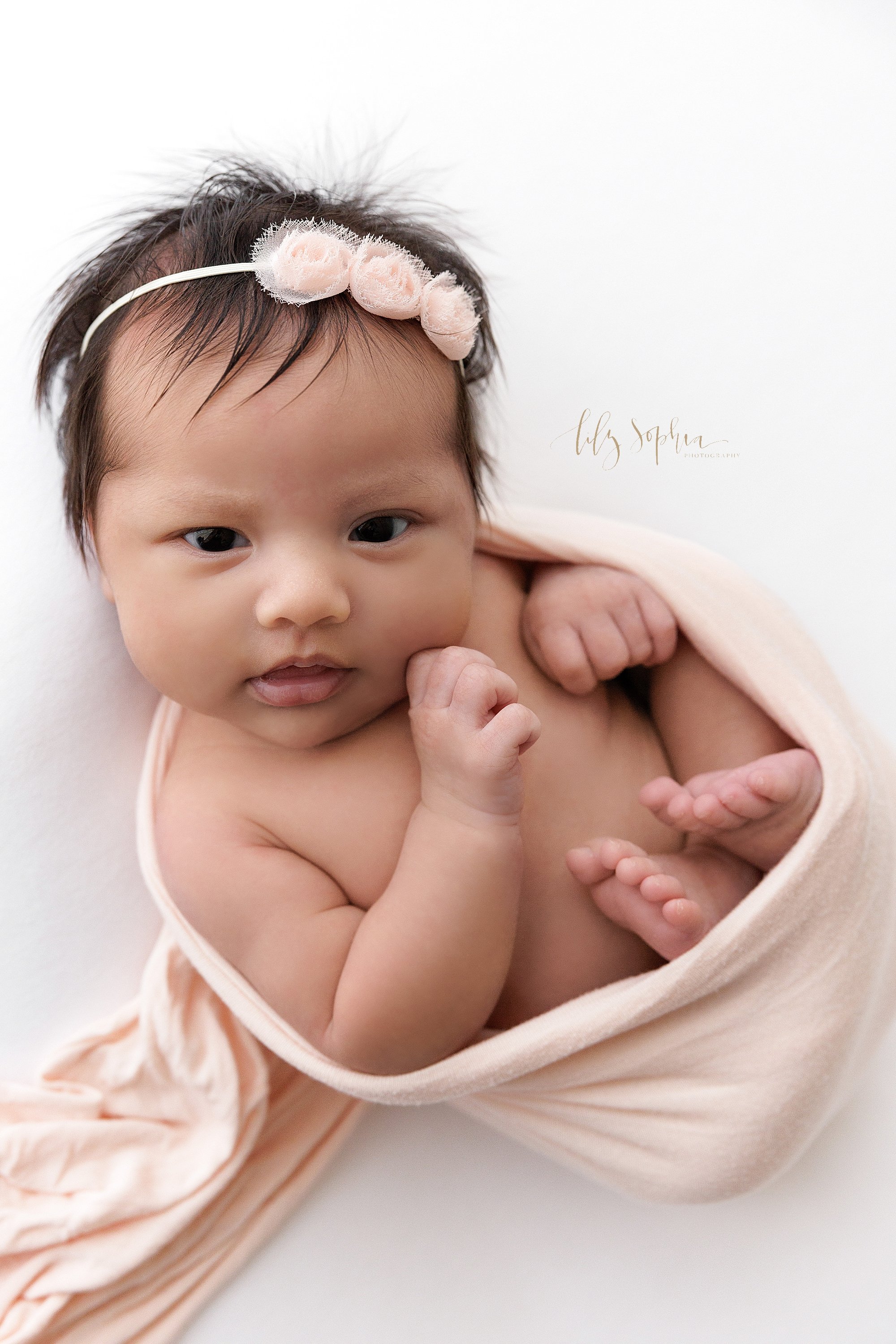  Newborn photo of an awake newborn baby girl cradled in a stretchy swaddle and wearing a delicate rose headband taken in a natural light studio near Old Fourth Ward in Atlanta. 