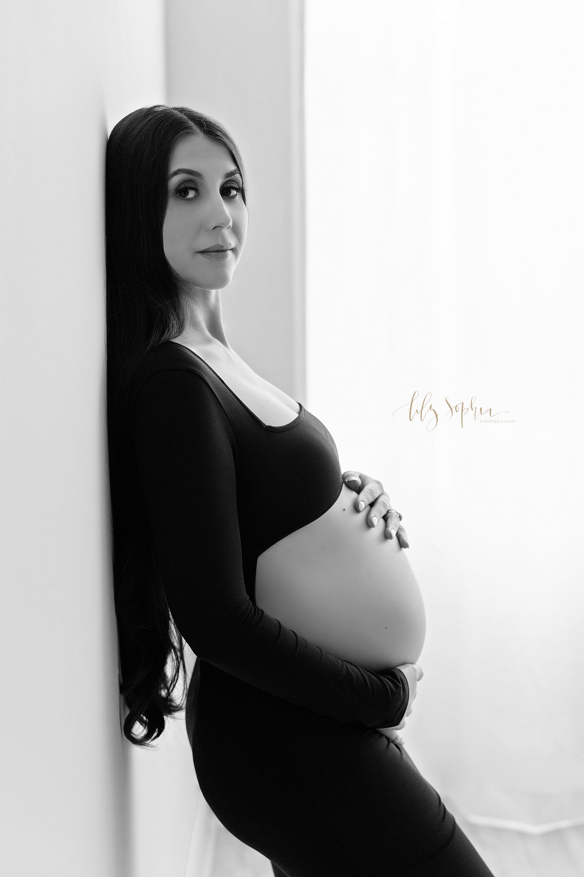  Modern maternity photo session of a pregnant mother as she stands against a wall with her hands framing her bare belly as she stands next to a window streaming natural light in a photography studio near Midtown in Atlanta, Georgia. 