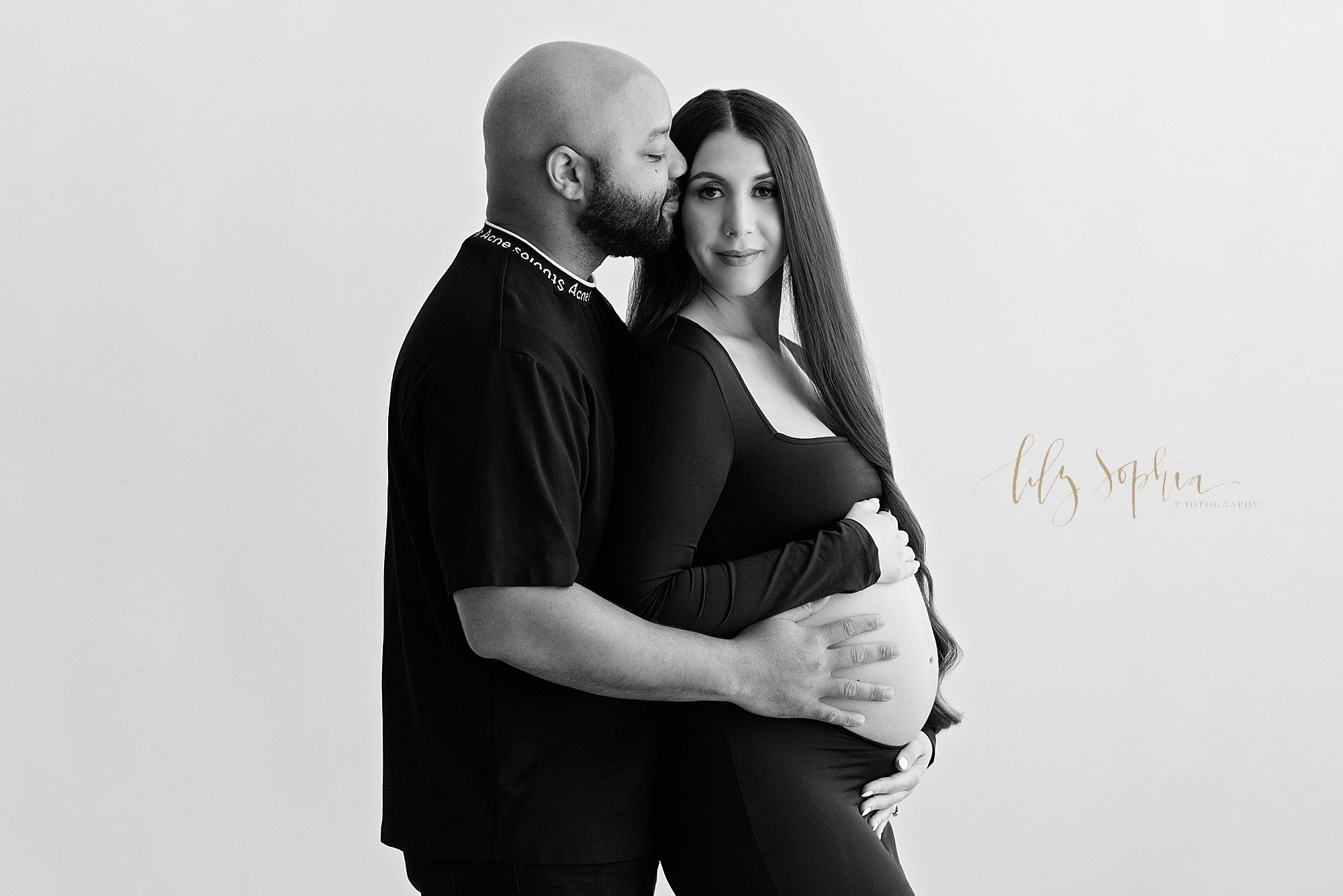  Black and white maternity portrait of a pregnant mother baring her womb in a jersey knit dress as she frames her womb and her husband stands behind her with his right hand on her womb and kissing his wife’s head taken in a natural light studio near 
