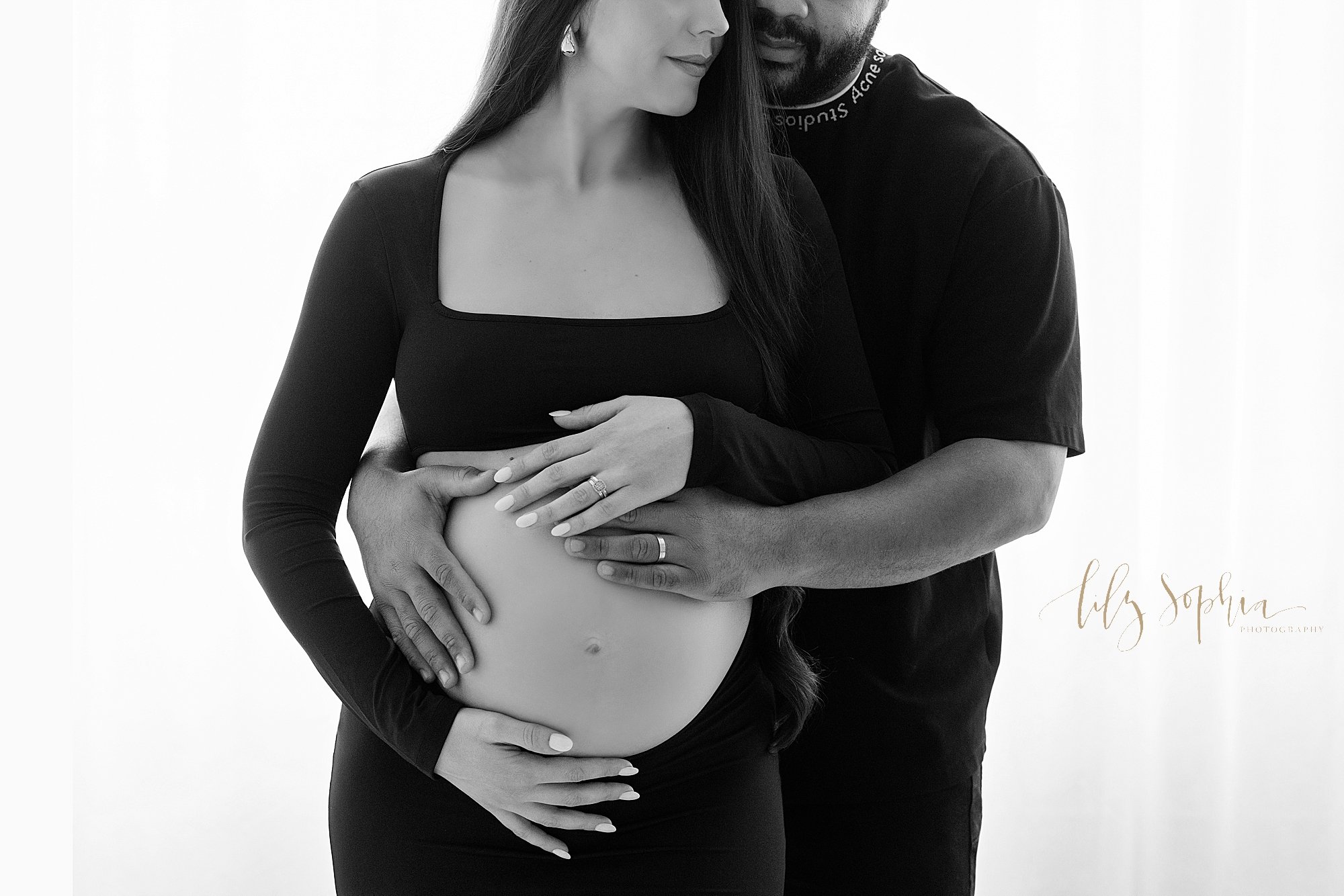  Maternity photo in black and white of a pregnant mother baring her belly in a jersey knit dress and framing her belly as her husband stands behind her and places his hands on their child in utero taken in front of window streaming natural light near