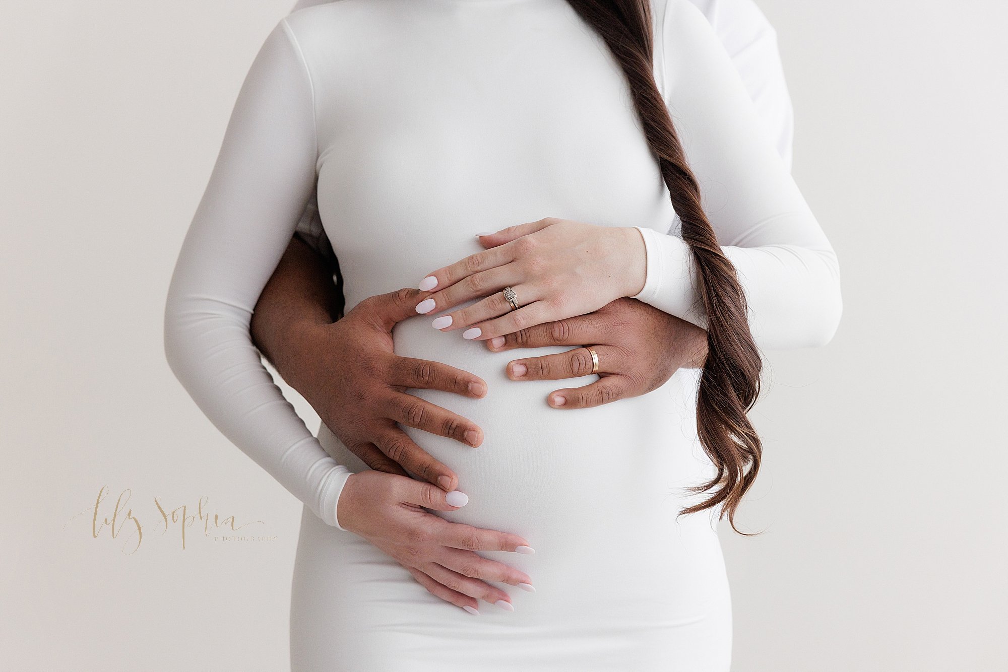  Close-up maternity portrait of a pregnant mother’s womb with mom’s hands framing her belly and her husband’s hands wrapped from behind and holding their child in utero taken in Ponce City Market in Atlanta at the Lily Sophia Photography Studio. 