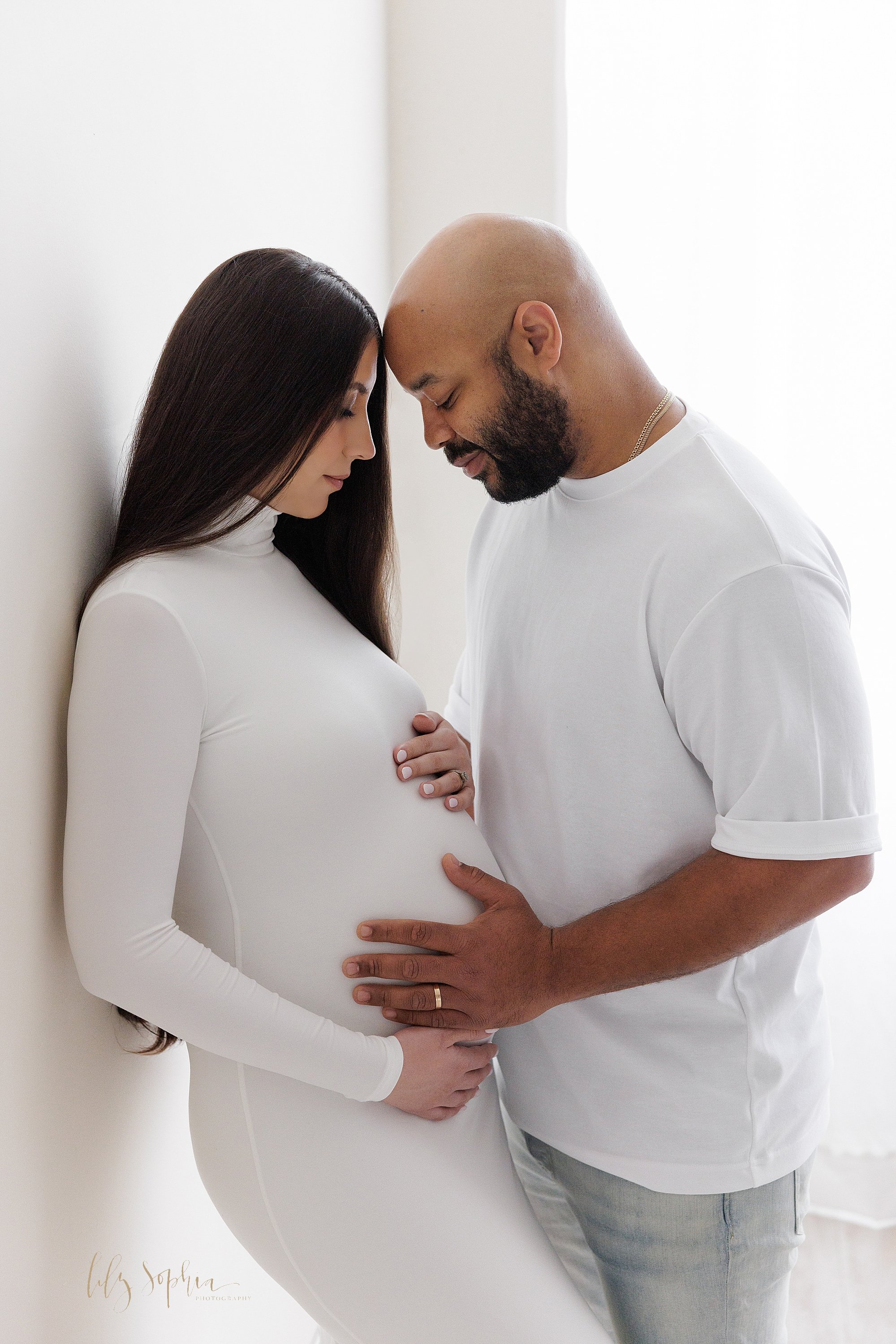  Maternity photo shoot of an expectant mother wearing a full-length knit jersey white long-sleeved gown with her hands framing her belly as her husband faces her wearing a white t-shirt and places his forehead against his wife’s forehead and his left