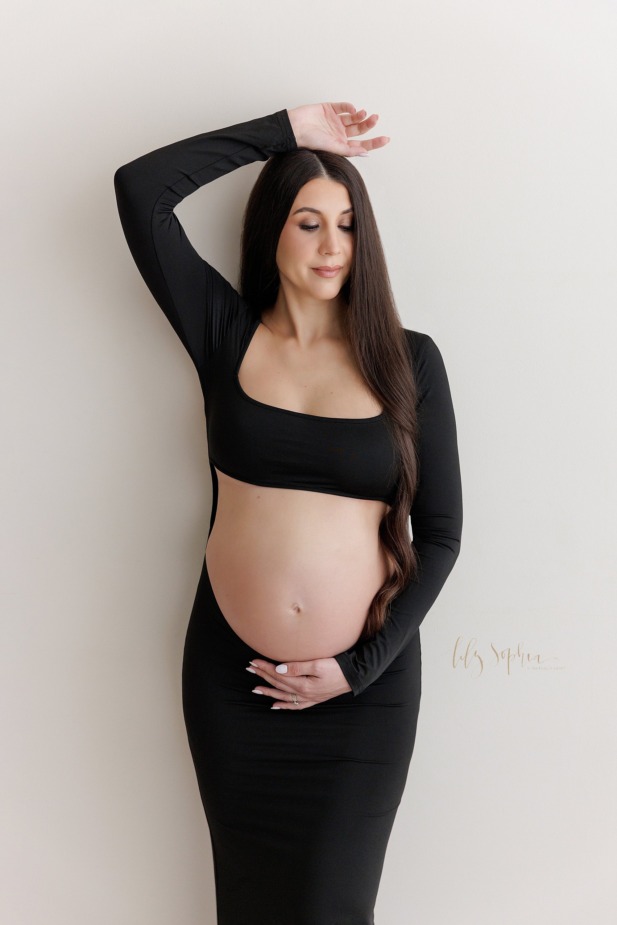  Modern maternity portrait of a pregnant woman wearing a jersey knit belly baring gown as she stands against a wall with her right arm bend above her head and her left hand holding the base of her belly taken in a studio near Oakhurst in Atlanta that
