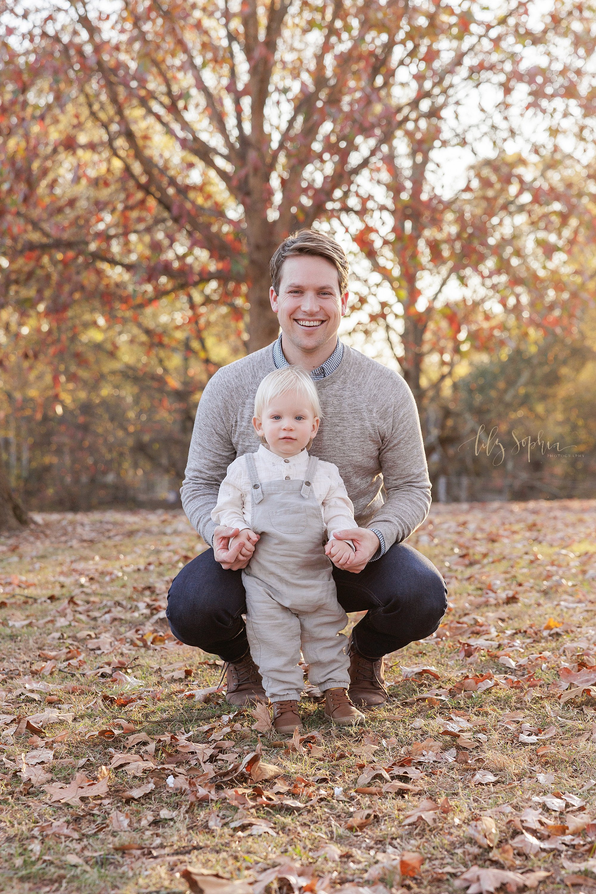  Father and son portrait with dad squatting behind his toddler son and holding his hands in a park carpeted with autumn leaves near Atlanta, Georgia at sunset. 