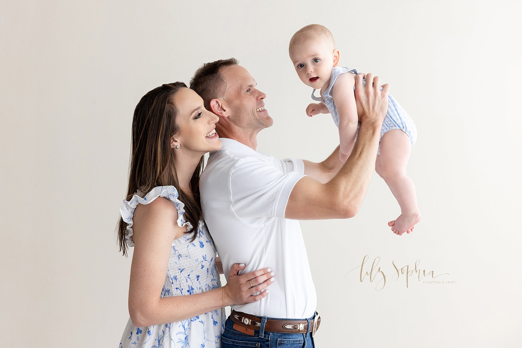  Family baby photo of a father holding his baby boy above his head as his wife stands behind him looking over his shoulder taken in a natural light studio near Ansley Park in Atlanta. 