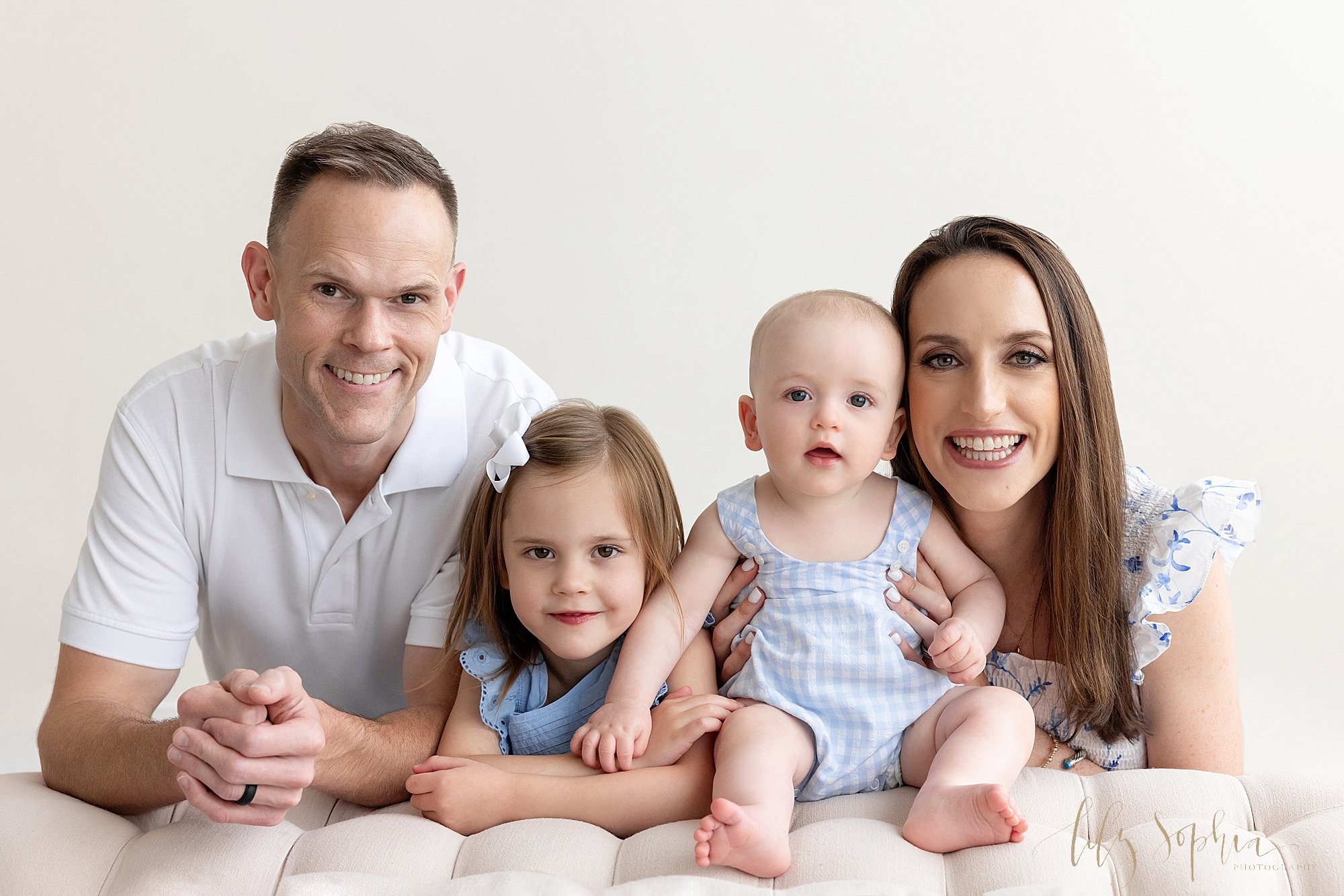  Family portrait of a father kneeling behind a tufted bench with his young daughter next to him, his baby boy sitting on the bench and his wife holding the baby boy taken next to a window streaming natural light near Alpharetta in Atlanta in a photog