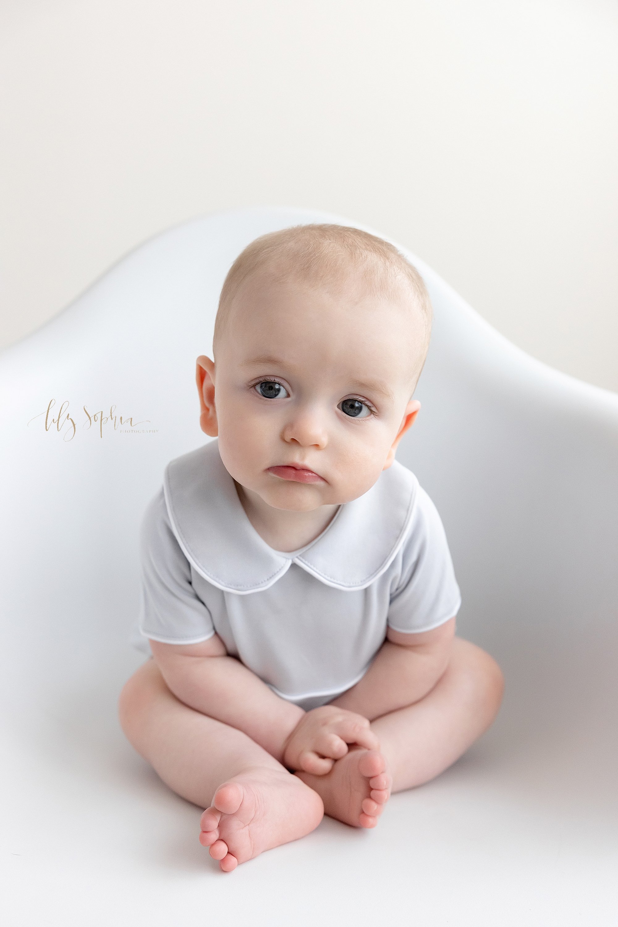  Sitting milestone portrait of a baby boy as he sits in a white molded chair in a studio using natural light near Poncey Highlands in Atlanta, Georgia. 