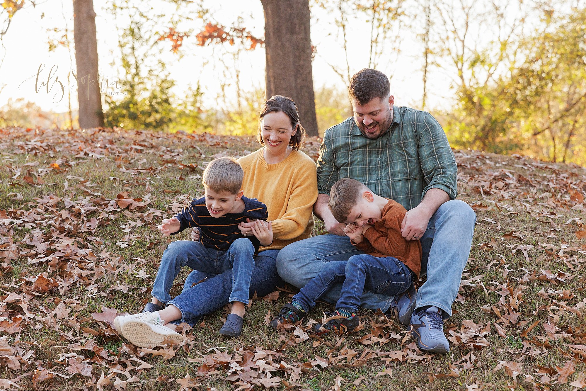  Fun natural family photo of a mother and father sitting on the ground among the fallen leaves in a park at sunset as each parent holds one of their rambunctious sons on their laps taken near Atlanta. 