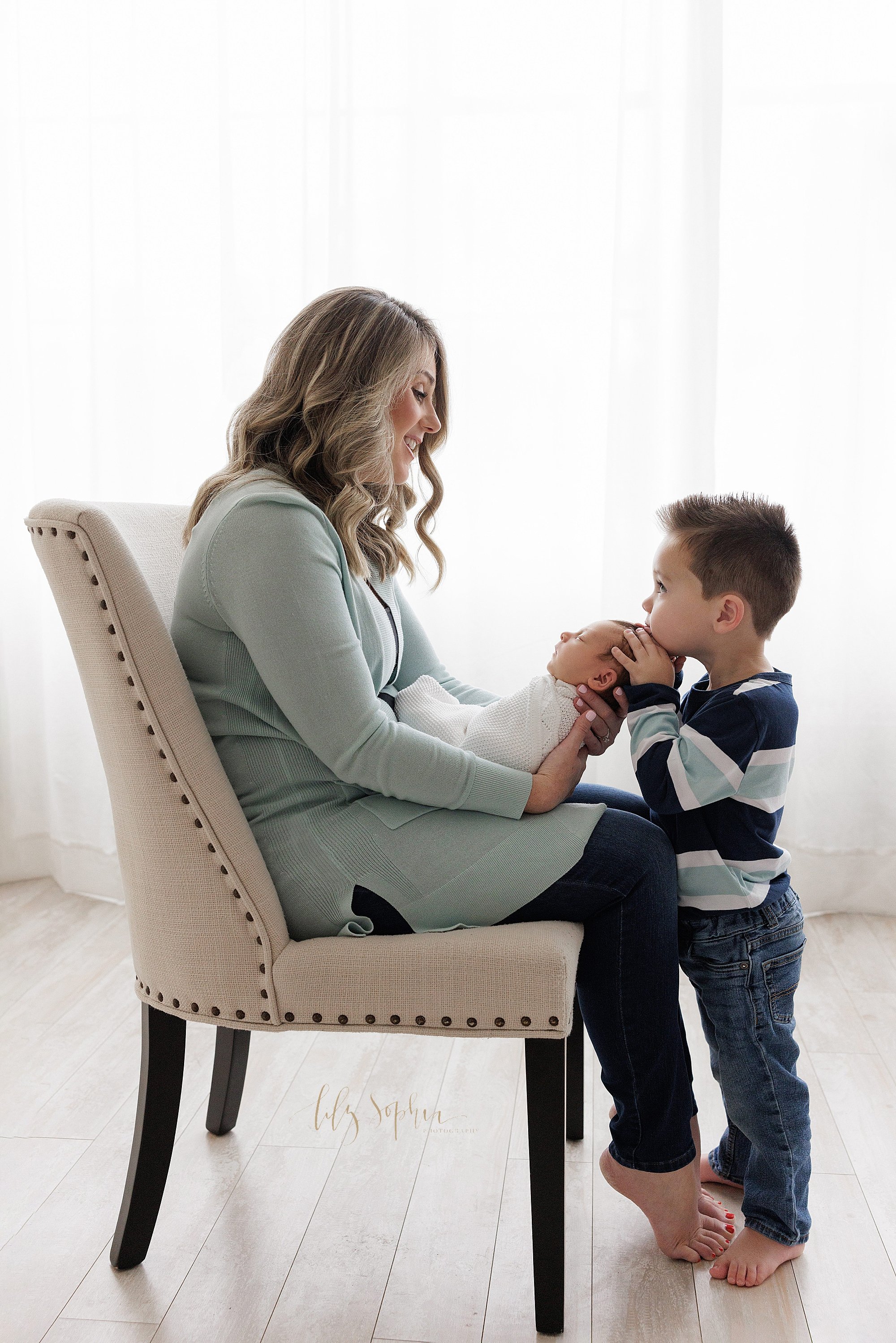  Newborn family portrait of a mother sitting in an upholstered chair with her newborn son on her lap as her young son stands facing her, holds his brother’s head in his hands and rests his head on the crown of the newborn boy taken in front of a wind