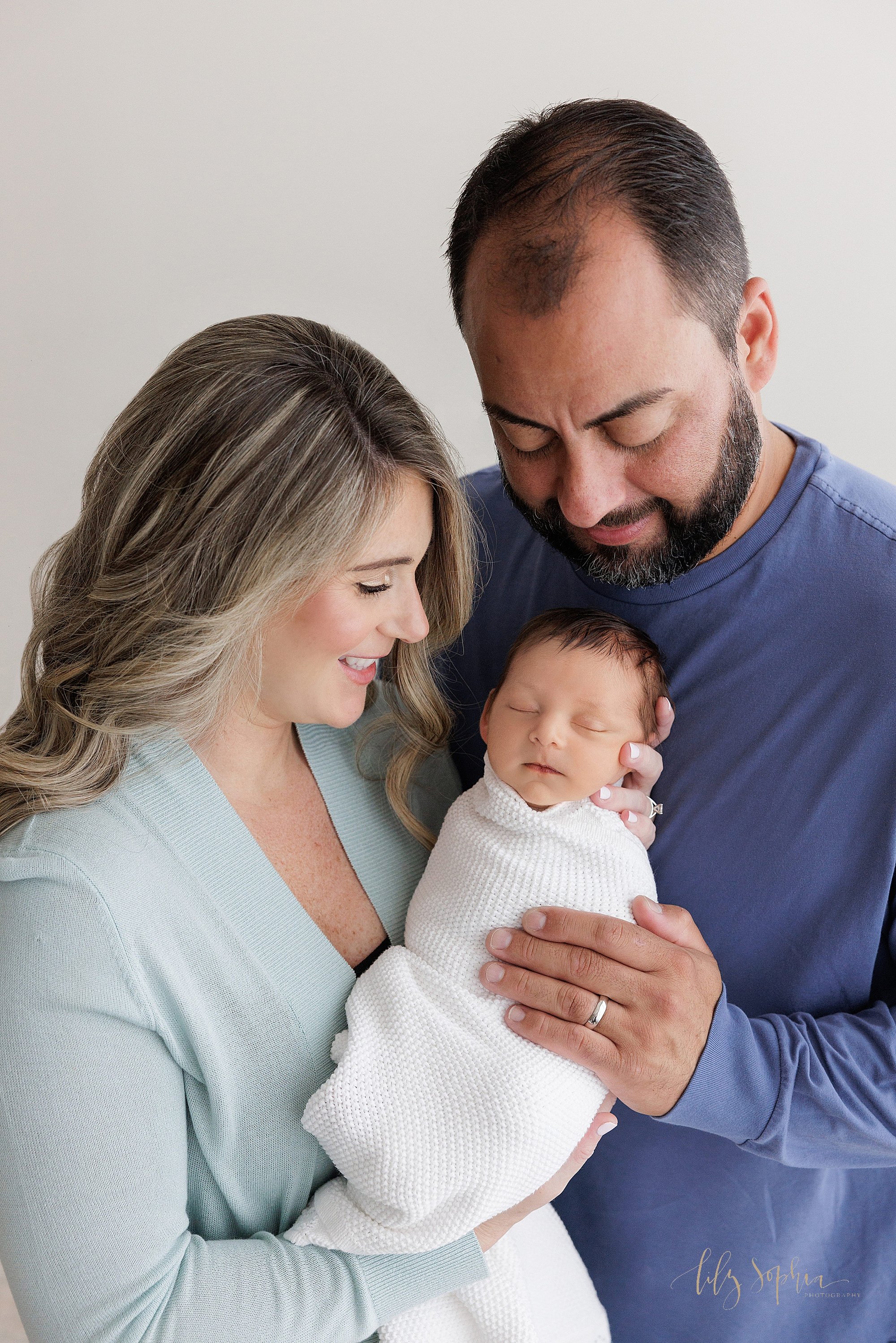  Newborn family portrait of a mother holding her peacefully sleeping newborn son in her arms as her husband stands next to her and places his left hand on his son and the parents admire him taken near Kirkwood in Atlanta in a natural light photograph