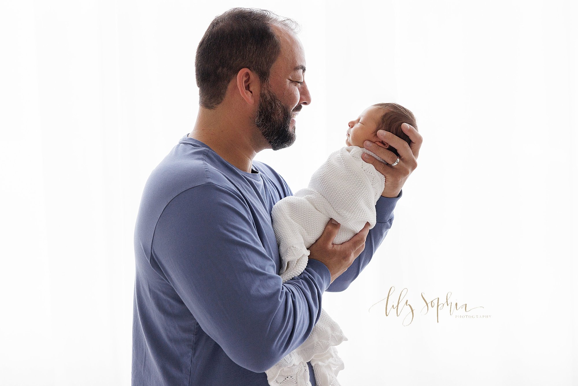  Newborn picture of a father holding his newborn baby boy in front of his chest and smiles as he talks to him while standing in front of a window streaming natural light into a photography studio near Oakhurst in Atlanta. 
