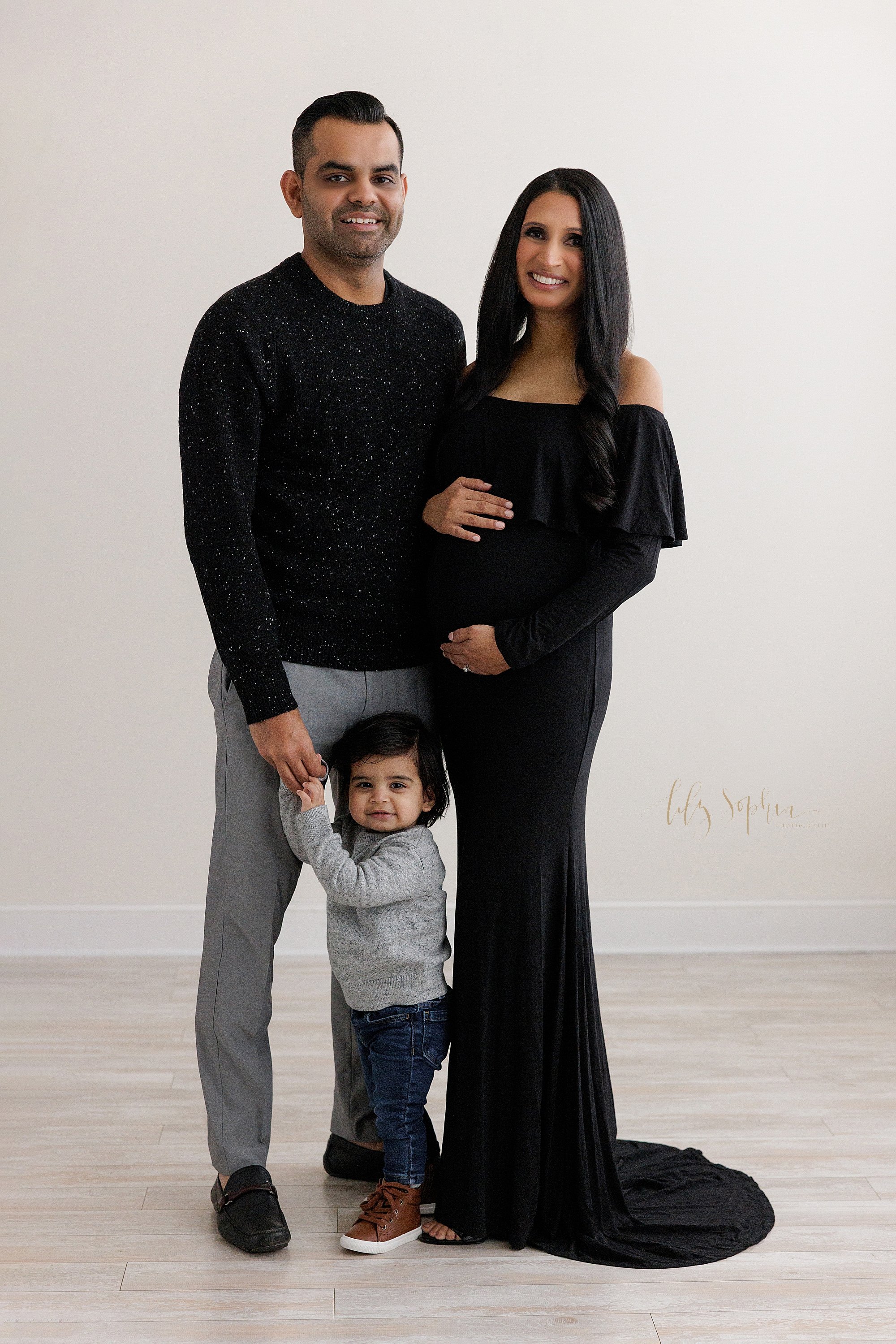  Maternity photo shoot of a pregnant mother wearing an off-the-shoulder black jersey full-length gown with her hands framing her child in utero as her husband stands to her right with their toddler son in front of him holding his hand taken in a natu