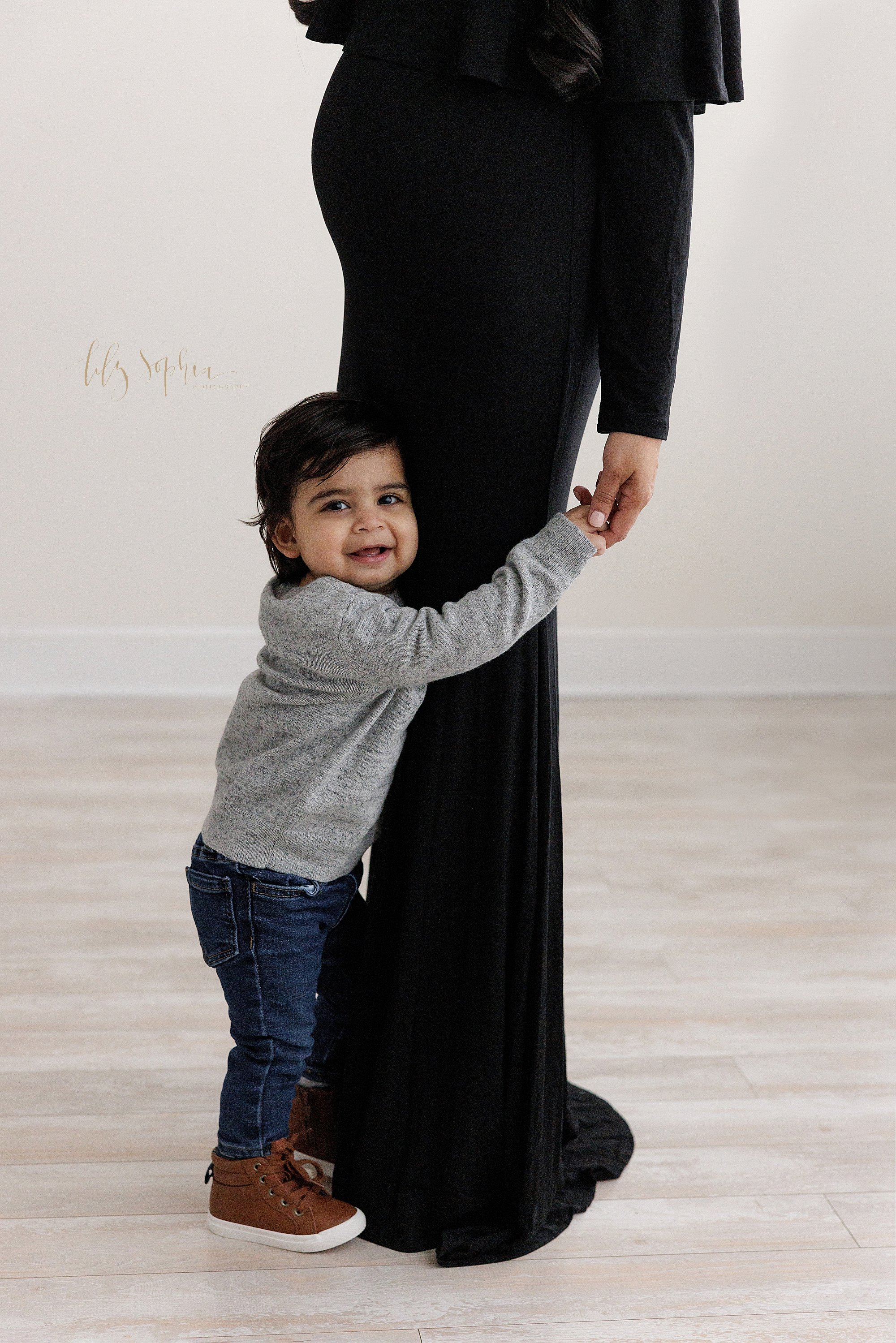  Family maternity portrait of a pregnant mother standing in a black jersey knit full-length gown in a photography studio with her toddler son facing her and hugging her legs with his left hand and holding his mother’s left hand with his right hand ta