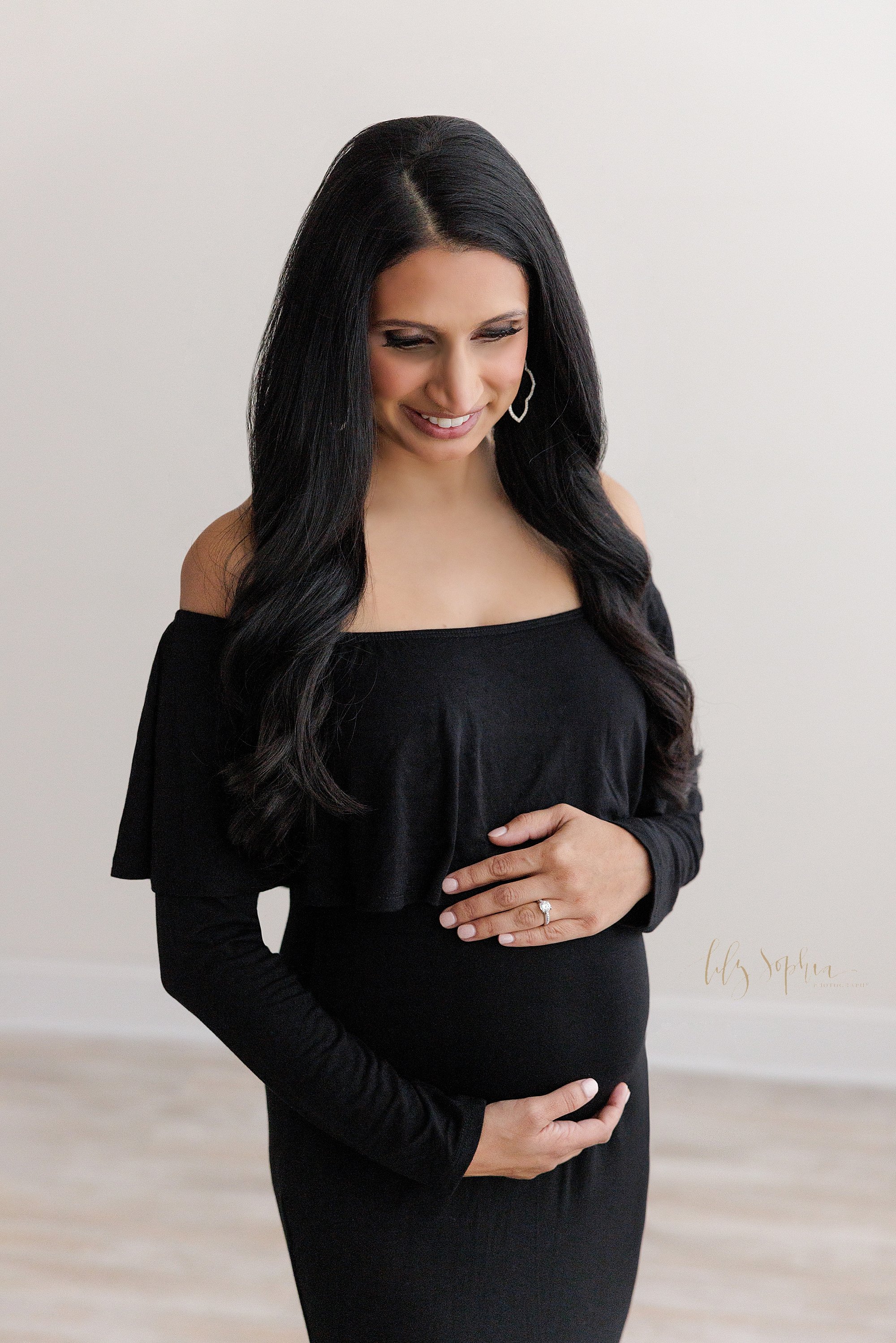  Maternity picture of an expectant mother wearing and off the shoulders long-sleeved black jersey knit full-length gown as she stands in natural light in a studio framing her belly with her hands taken near Vinings in Atlanta. 