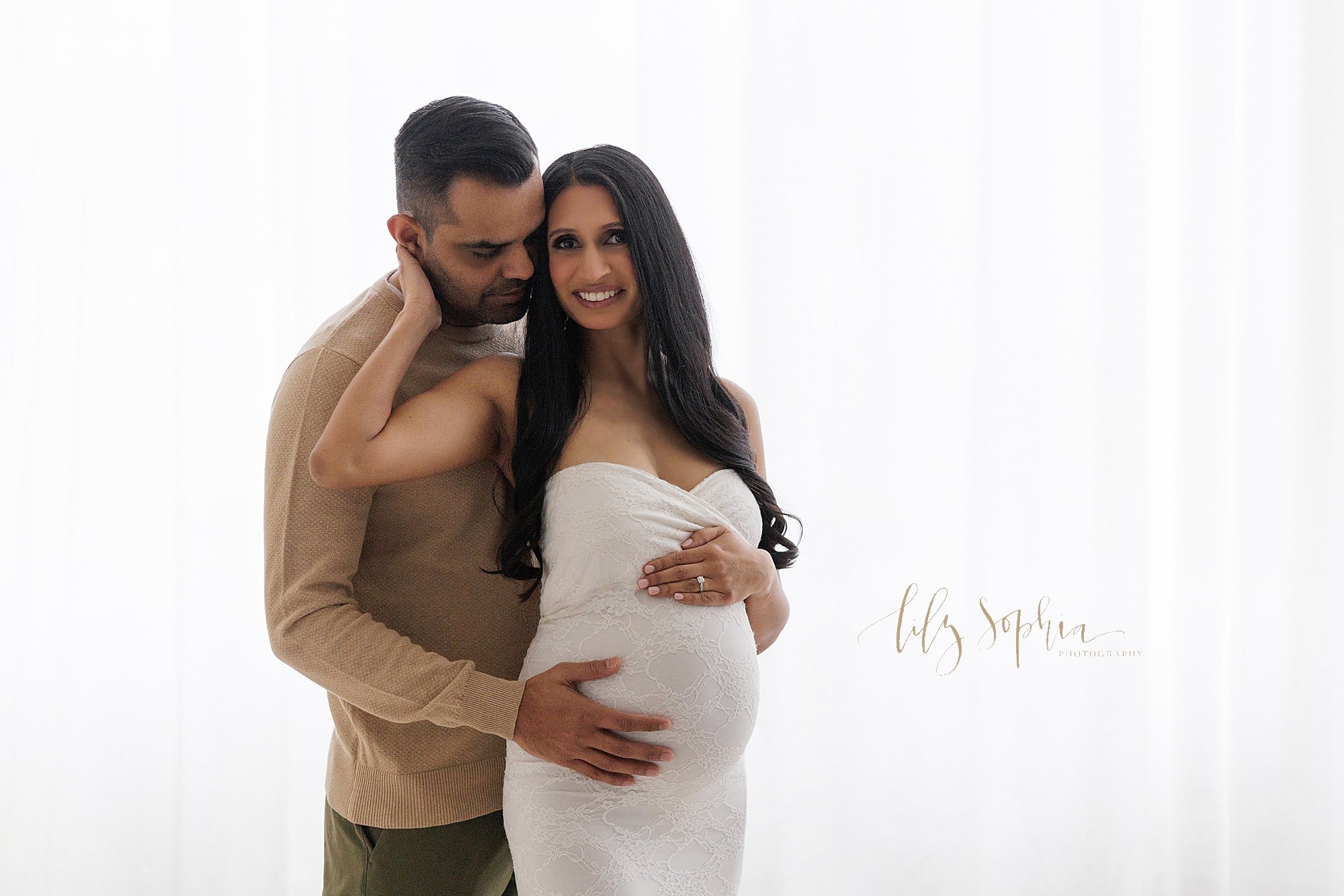  Maternity photo of a pregnant mother wearing a strapless white knit gown with a lace skirt standing in front of a window streaming natural light and placing her left hand on top of her belly and her right hand on the neck of her husband who stands b