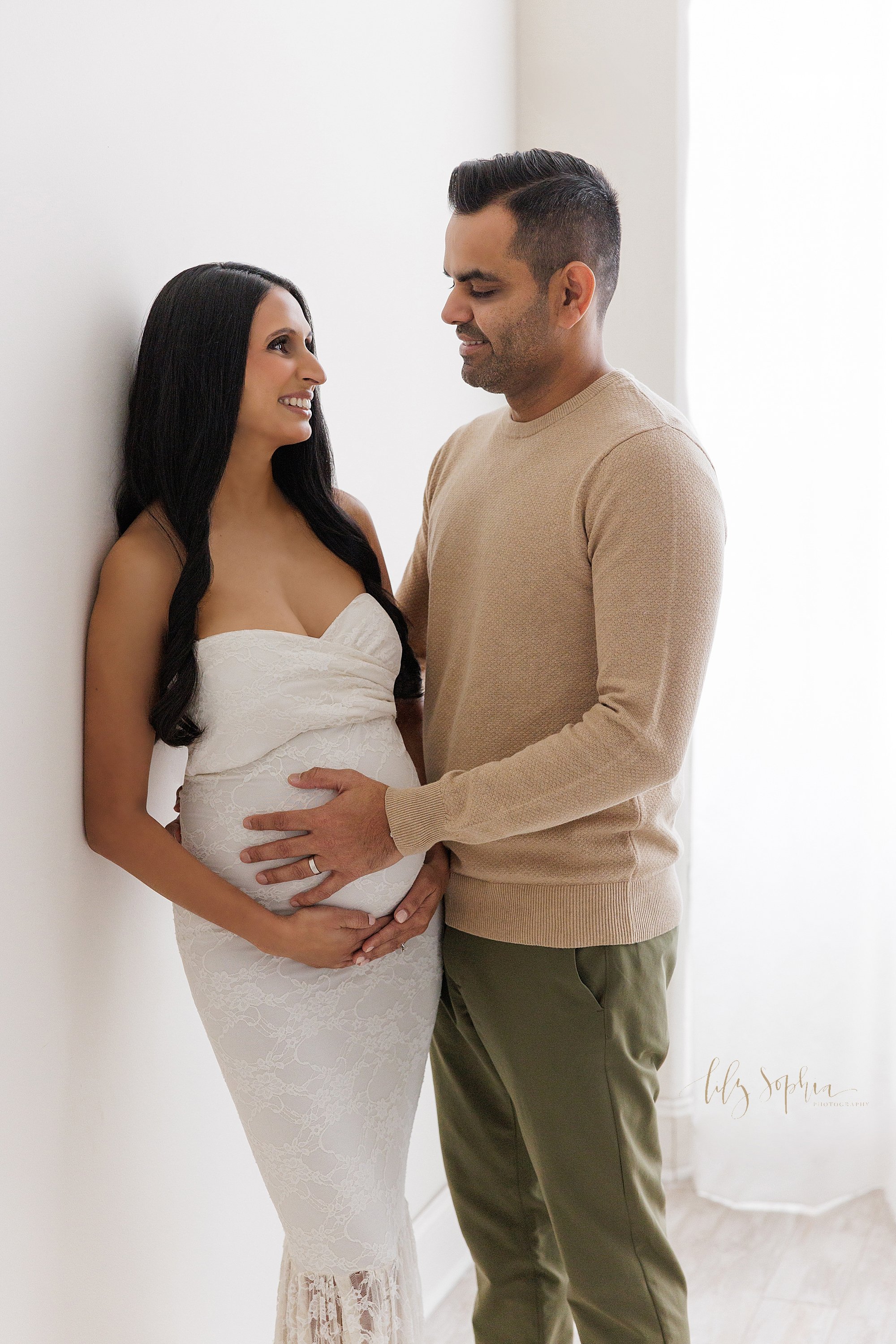  Maternity picture of a pregnant mother wearing a white strapless knit gown as she stands with her right shoulder against a wall and holding the base of her belly as her husband stands facing her and placing his left hand on their child in utero whil