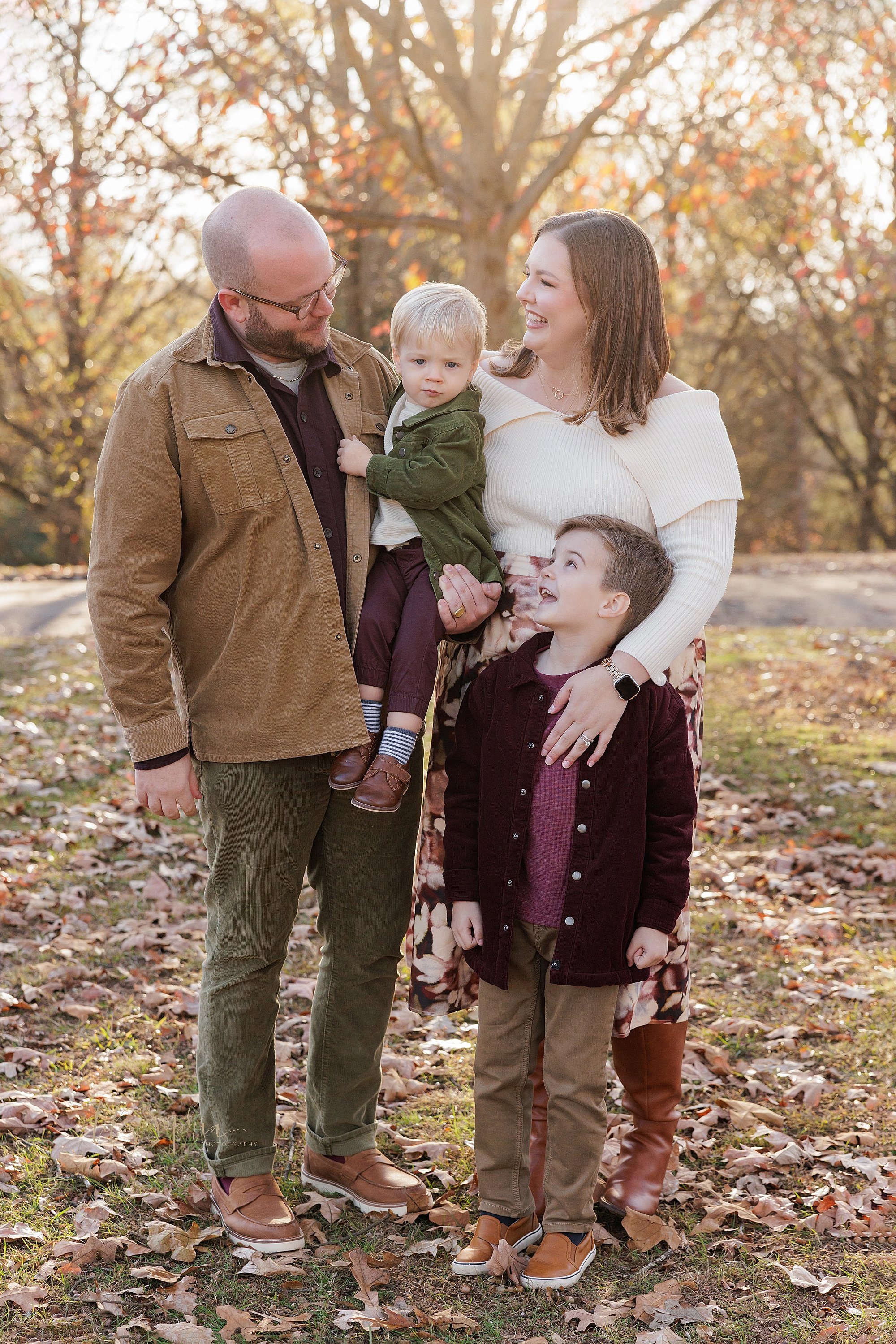  Family portrait of dad holding his toddler son in his right arm as mom stands beside him with their young son in front of her as she places her left hand on his shoulder and the family talks to one another during autumn in a park near Atlanta at sun