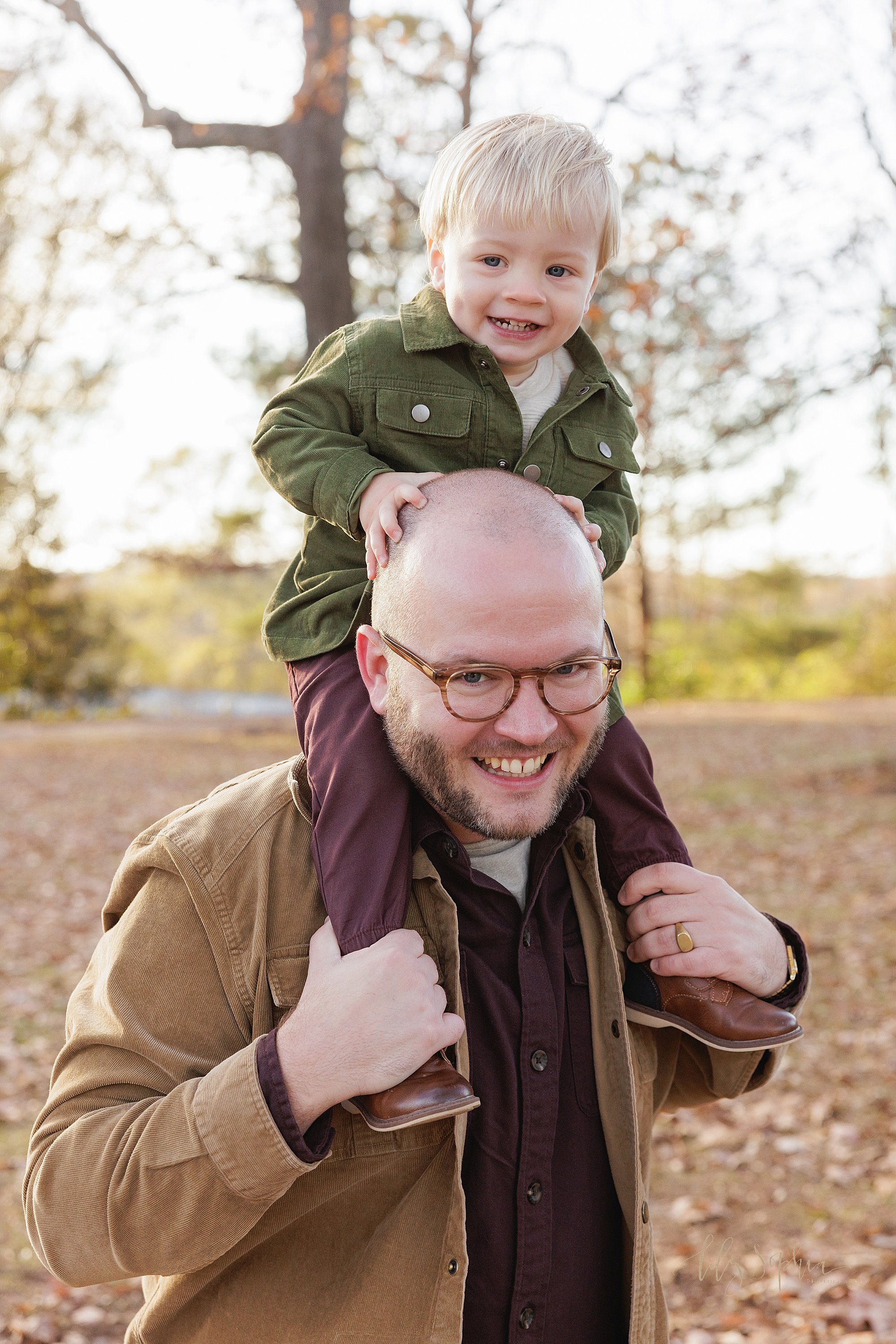  Father and son photo of a father holding his toddler son on his shoulders for a piggy back ride at sunset in an Atlanta park during autumn. 