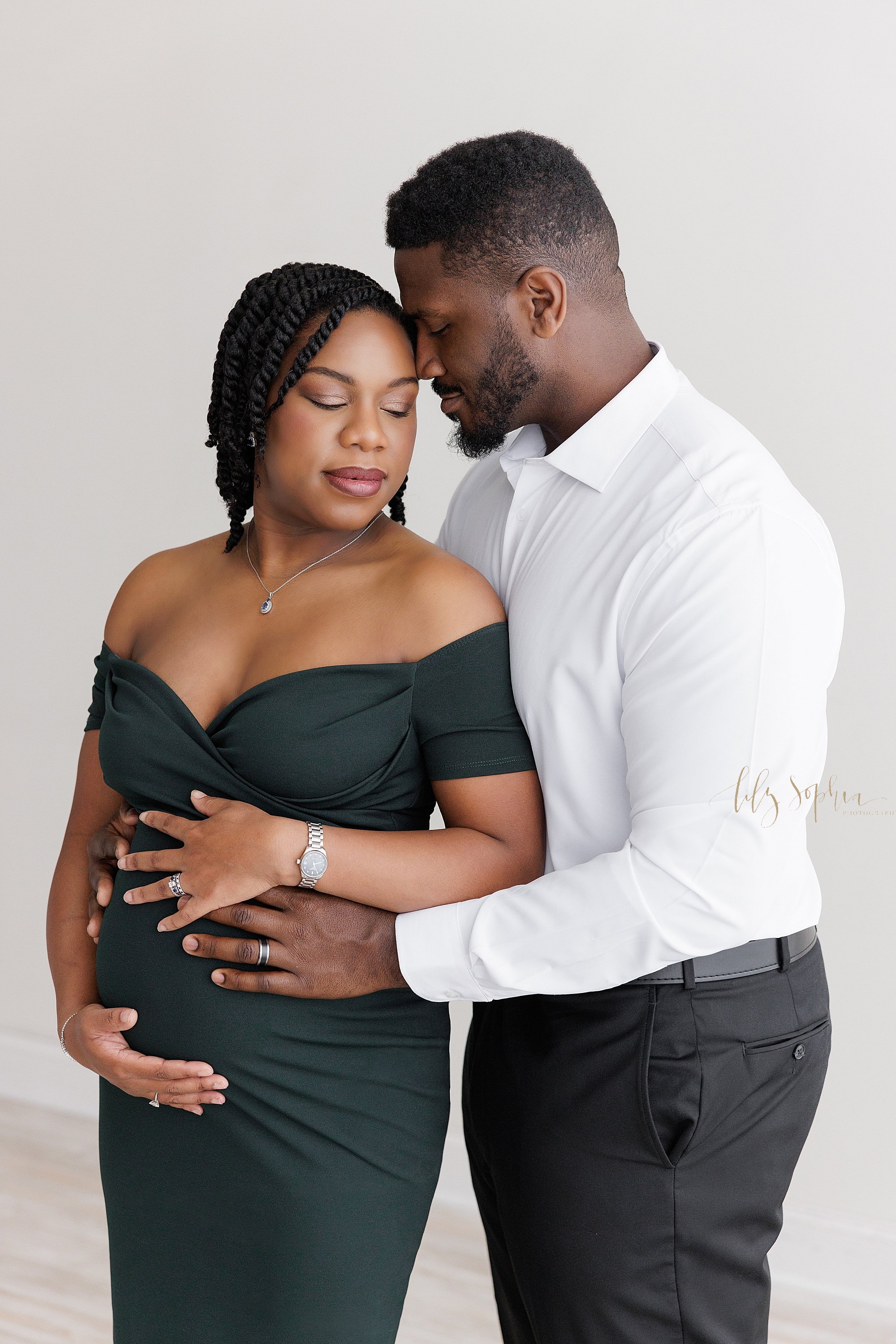  Maternity photo session of an African-American expectant couple as the husband stands next to his wife with his head against his wife’s head who is wearing an off the shoulder black gown as the two of them place their hands on their child in utero a