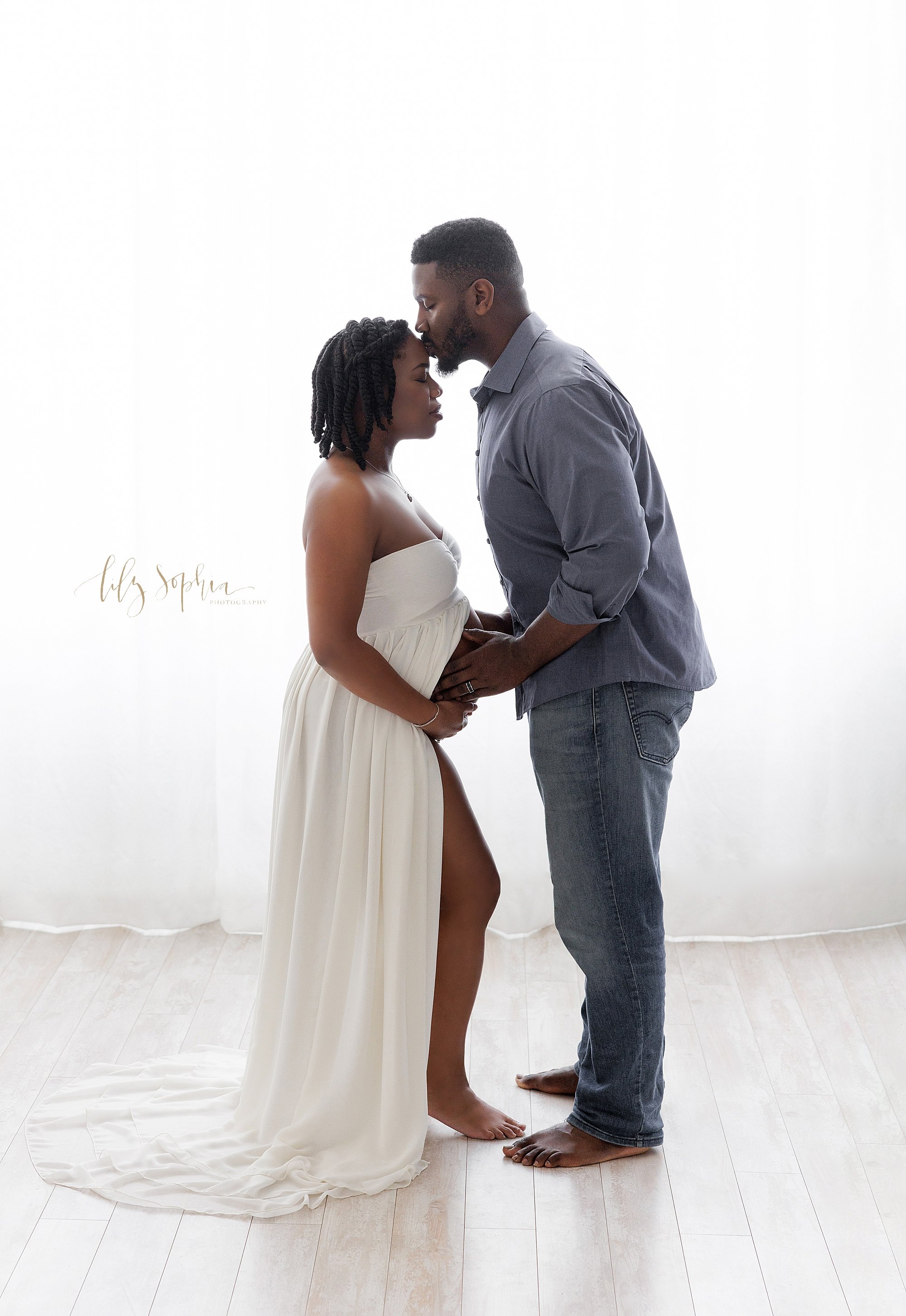  The love of an expectant African-American couple is captured in front of a window streaming natural light in a photography studio as the wife stands baring her belly in a white split front gown and her husband faces her with his hands on their child