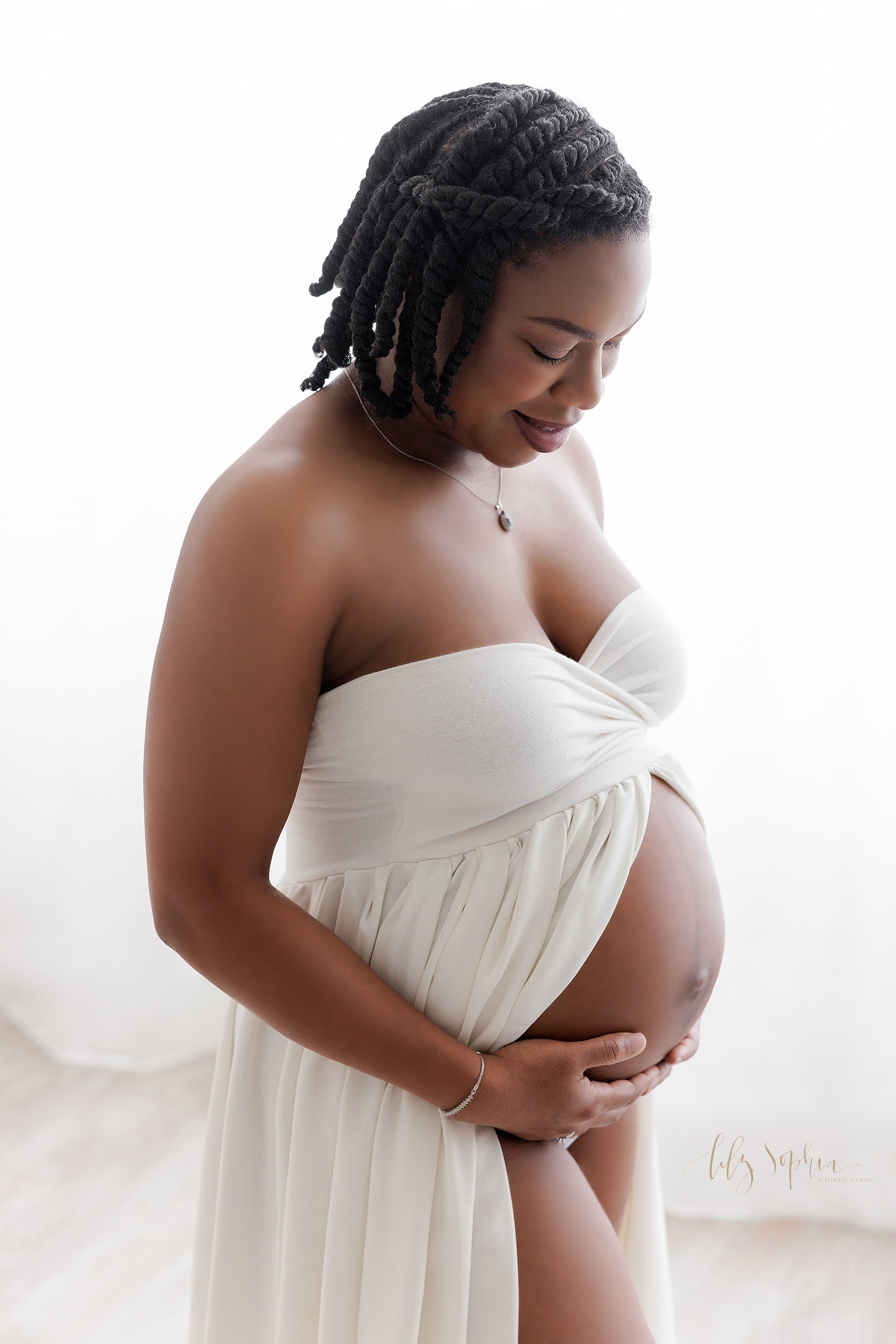  Maternity portrait of an expectant mother wearing a strapless split front white gown holding her bare belly as she stands in front of window streaming natural light in a photography studio near Decatur in Atlanta, Georgia. 