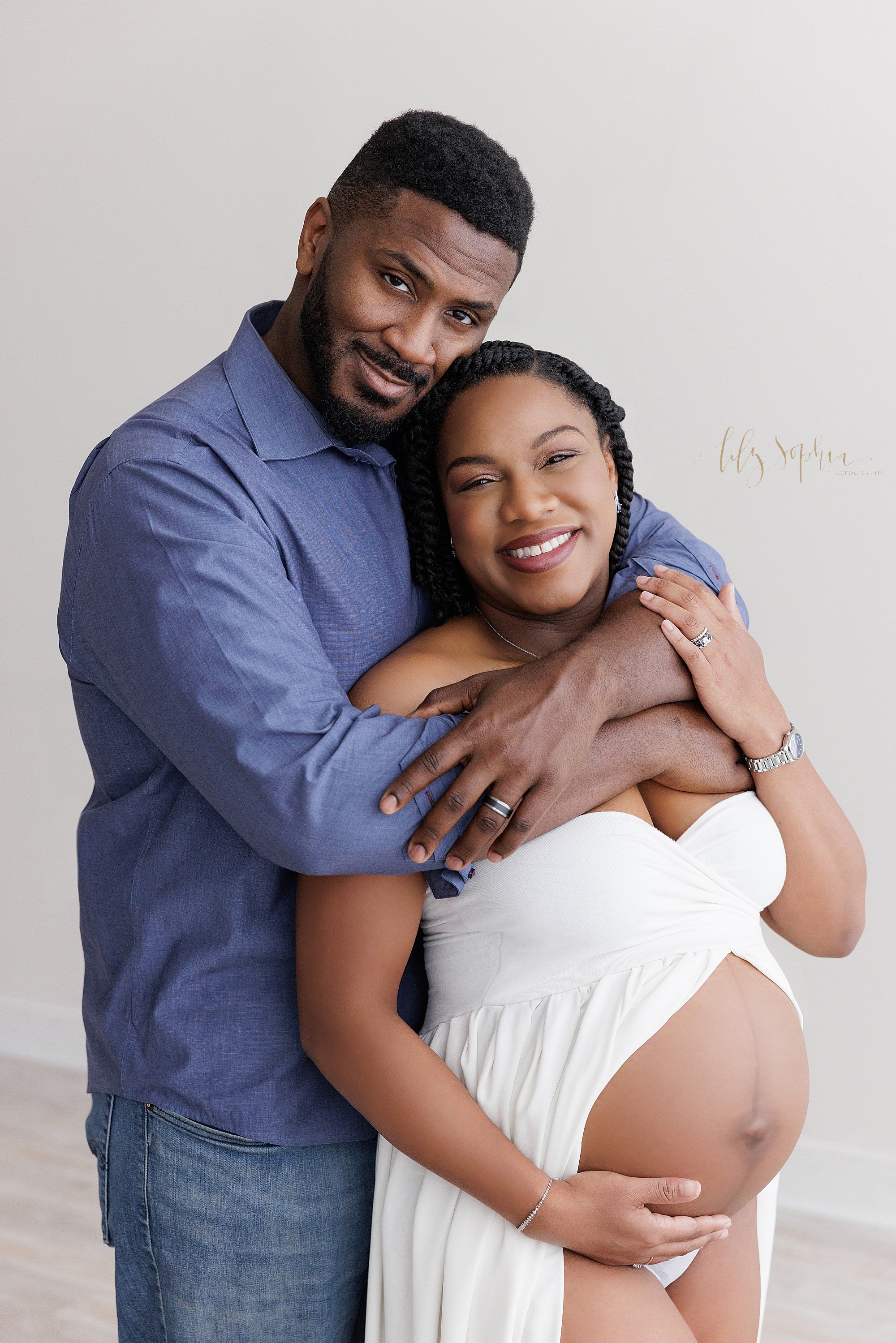  Maternity portrait of an African-American husband and wife as the husband stands with his wife’s back to his chest and wraps his arms around her with her belly bared as she wears a split-front gown taken next to a window streaming natural light near