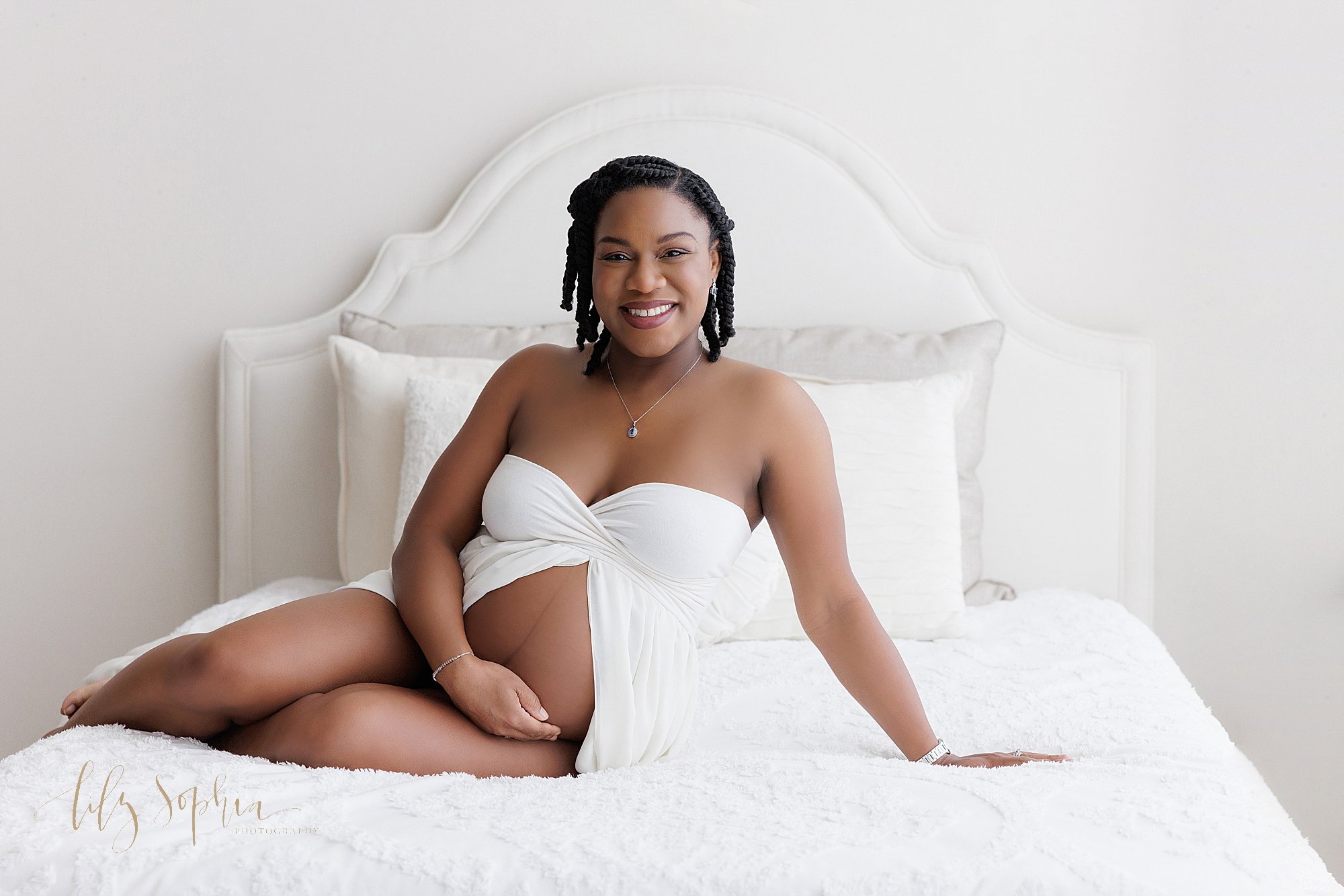  Maternity photo session of an African-American pregnant mother as she sits atop a bed wearing a strapless split front gown with her right hand holding her bare belly and her left hand supporting her on the bed taken in Ponce City Market in Atlanta i