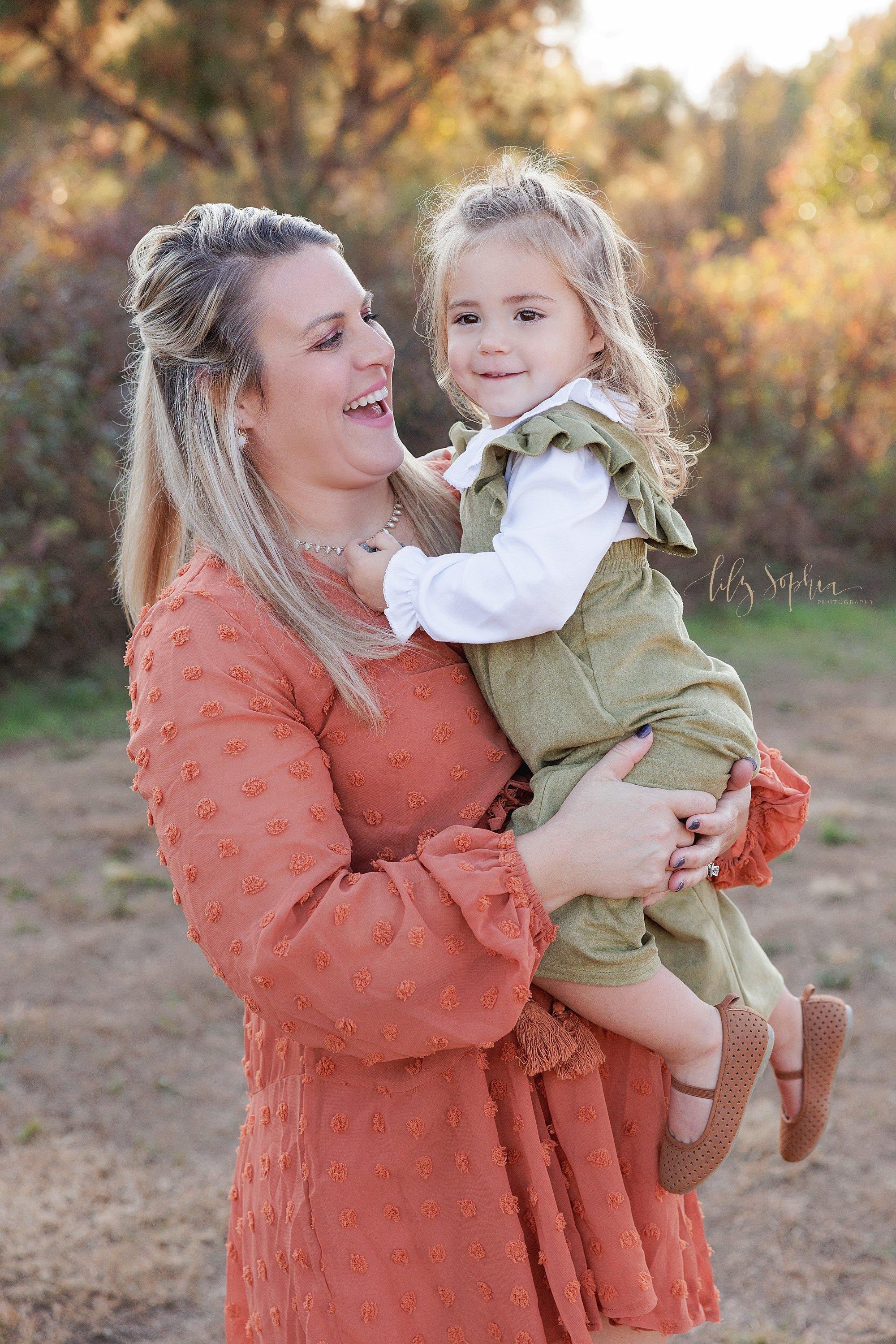  Family photo session in a field near Atlanta with a mother holding her young daughter as she plays with her mother’s necklace taken during autumn at sunset. 