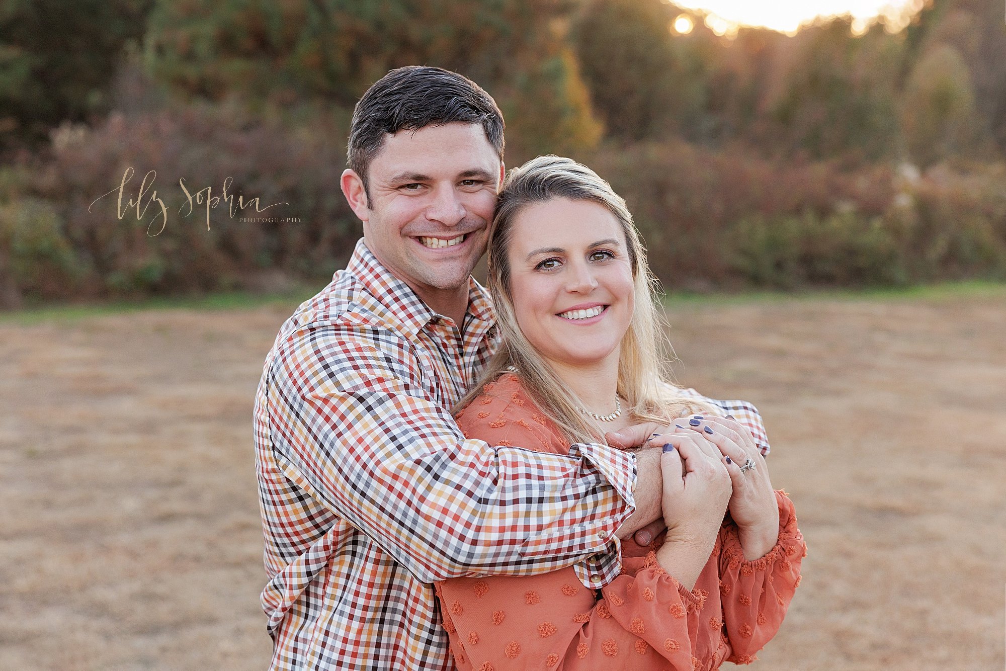  Family photo of a mom and dad as mom stands with her back against her husbands chest as he wraps his arms around her and she holds onto them taken in an Atlanta field during autumn at sunset. 