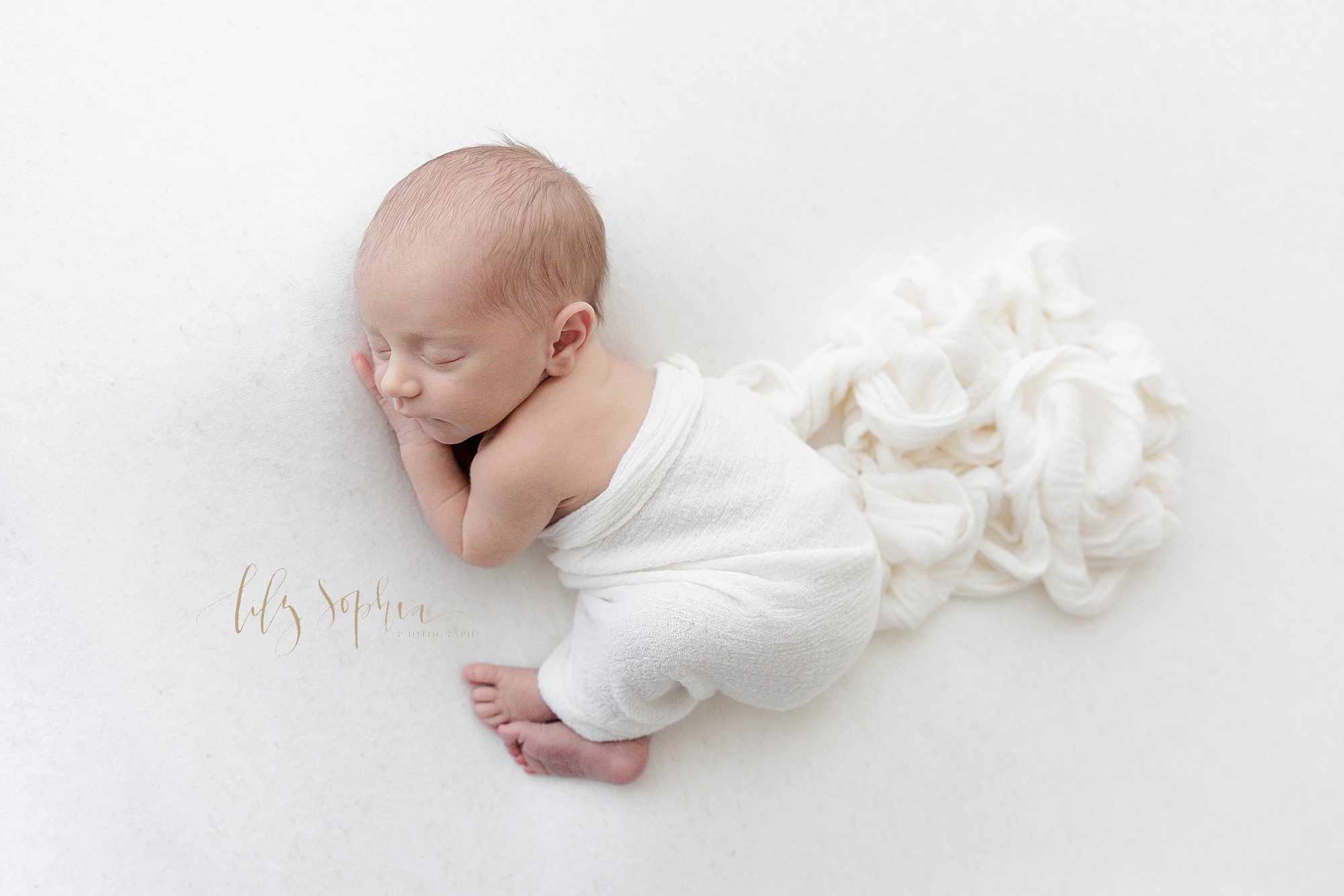  Newborn portrait of an infant boy as he sleeps on his side with his arms under his head and a soft white blanket drapes across him taken in a natural light studio near Virginia Highlands in Atlanta. 