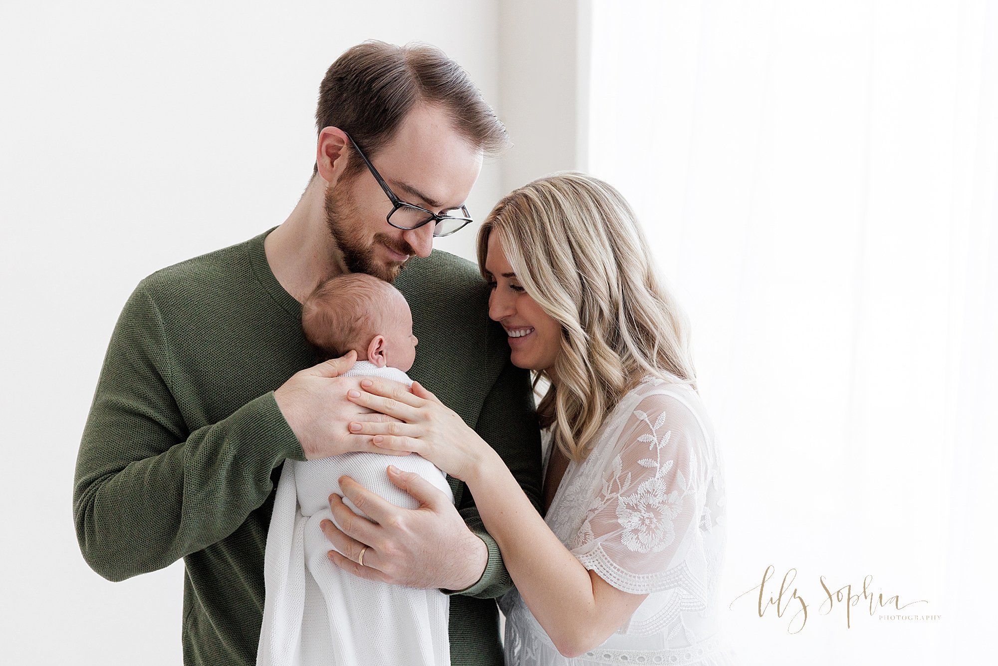  Newborn family portrait of a father standing next to a window streaming natural light while holding his newborn son against his chest with his wife standing next to him and the two of them admire their addition to their family taken near Kirkwood in