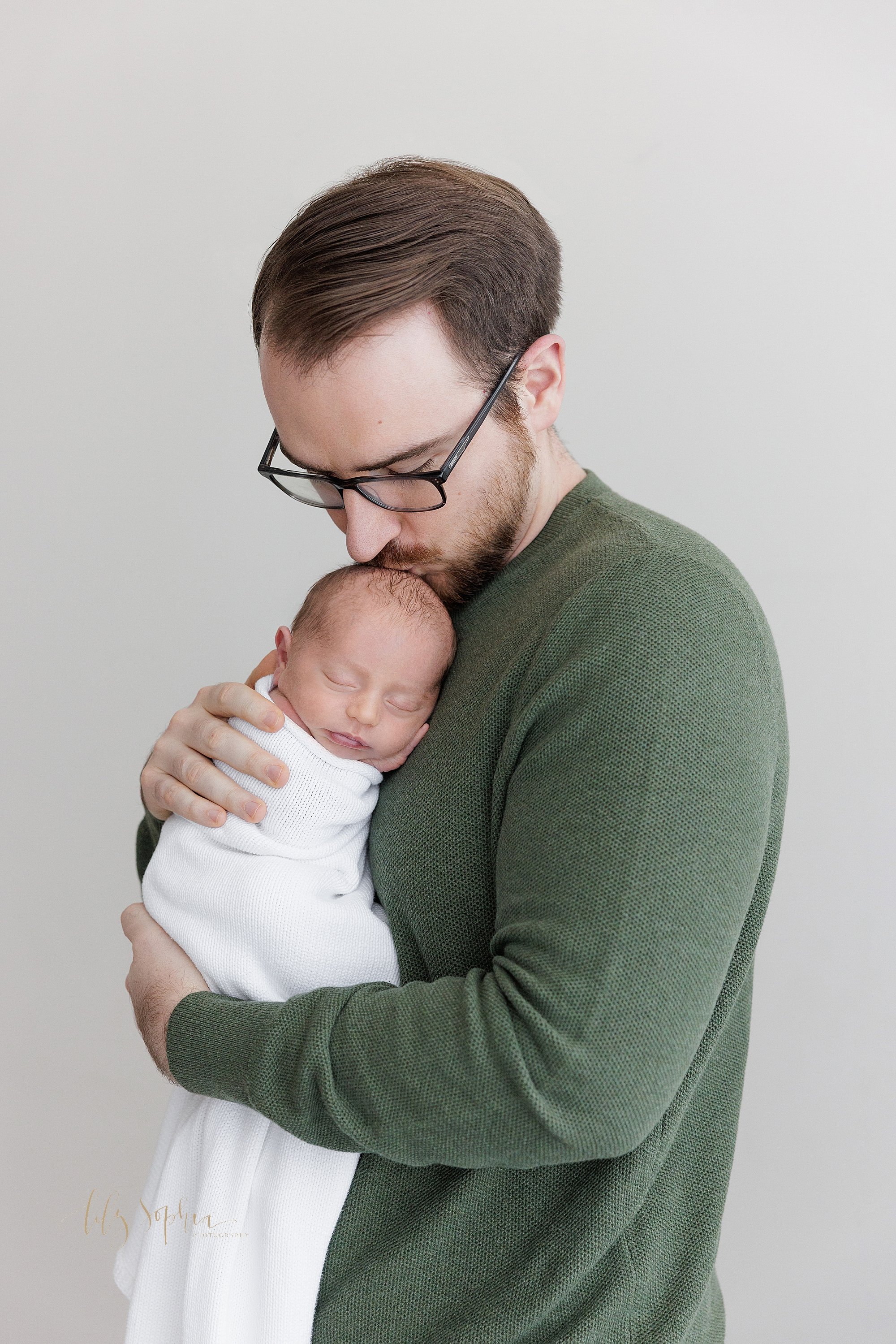  Newborn photo of a father’s love for his newborn son as he holds his peacefully sleeping infant boy against his chest and kisses the crown of his head while he stands in a studio using natural light near Sandy Springs in Atlanta, Georgia. 