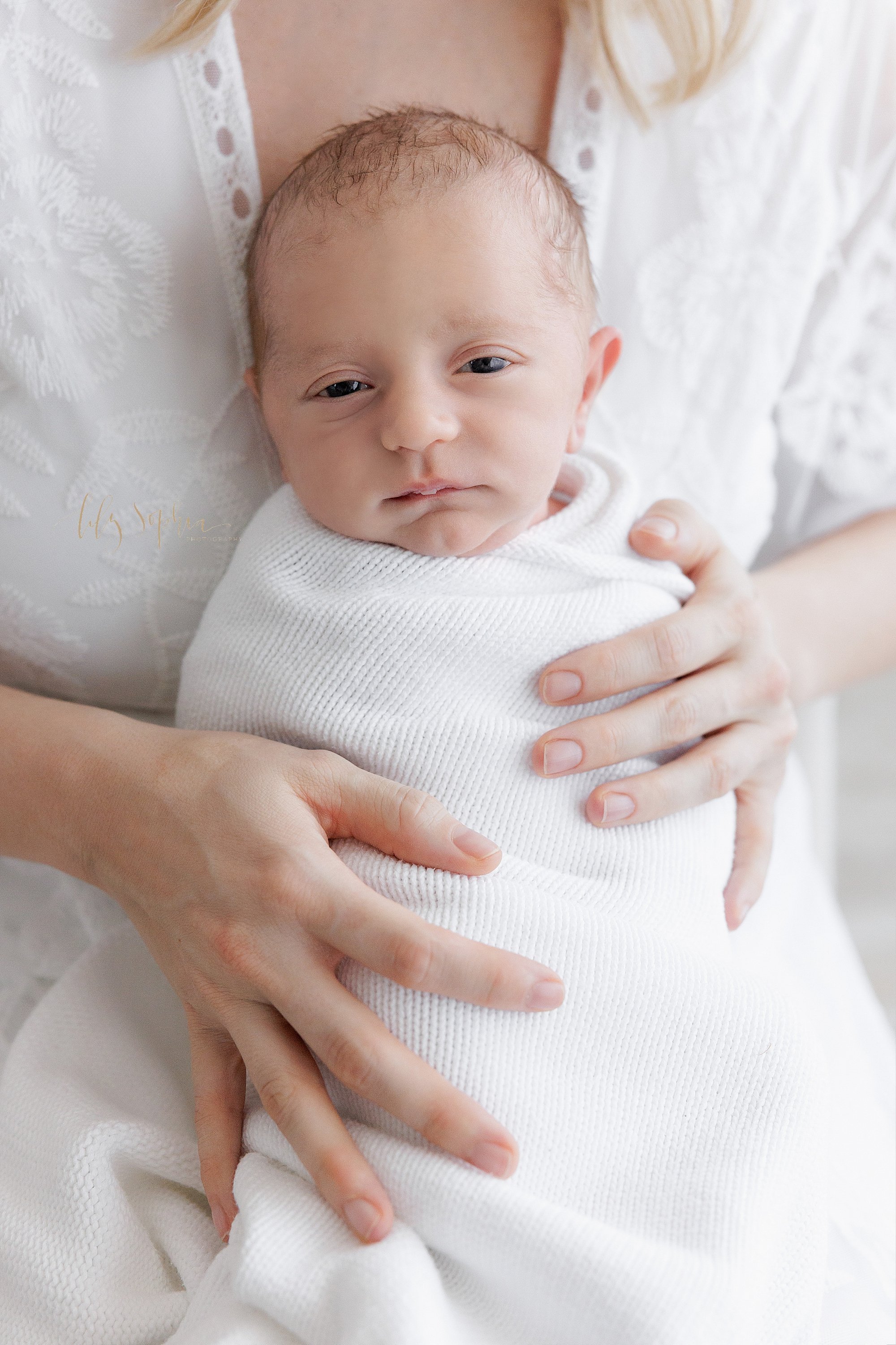  Newborn photo of an awake newborn baby boy as he is held in front of his mother as she sits in a chair with him swaddled to his chin in a soft white blanket taken near Brookhaven in Atlanta in a photography studio that uses natural light. 