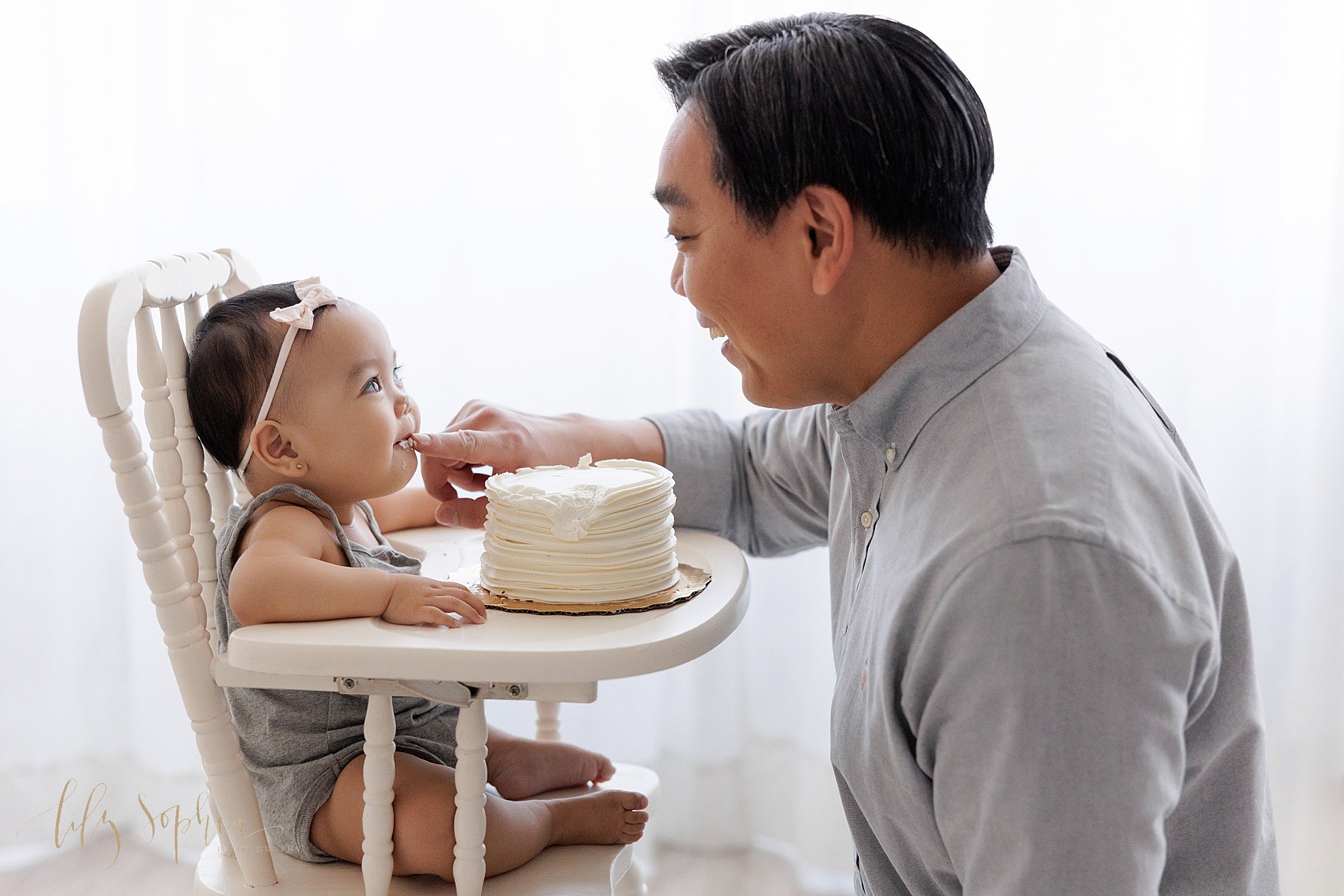  Smash cake first birthday photo of a one year old Asian girl sitting in an antique high chair with her father sitting in kneeling in front of her to give her some icing from the smash cake on the tray as the two look at each other taken in front of 