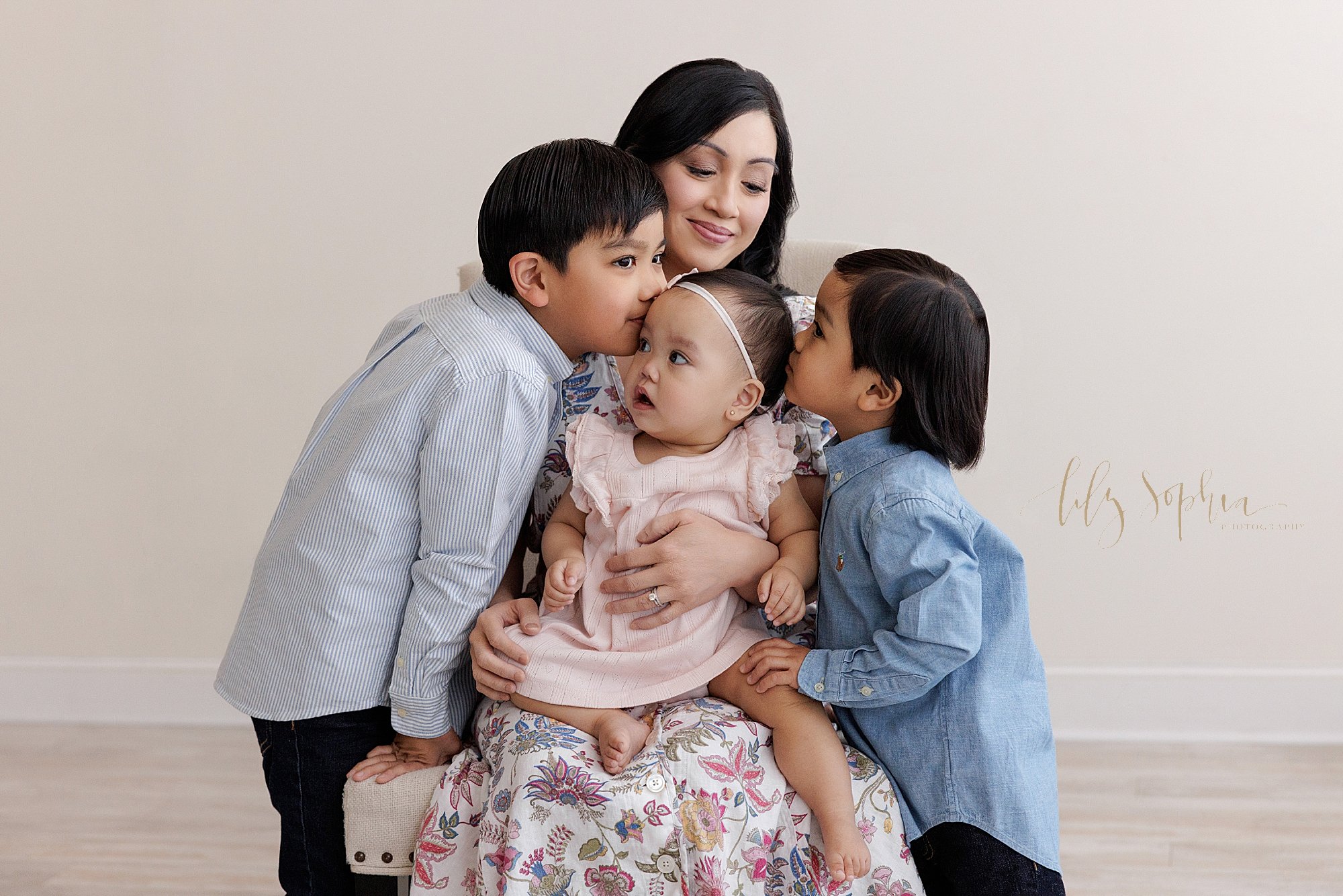  First birthday family photo of a mother holding her one year old baby girl on her lap as her sons stand on either side of her and kiss their sister’s head taken near Smyrna in Atlanta in a photography studio that uses natural light. 