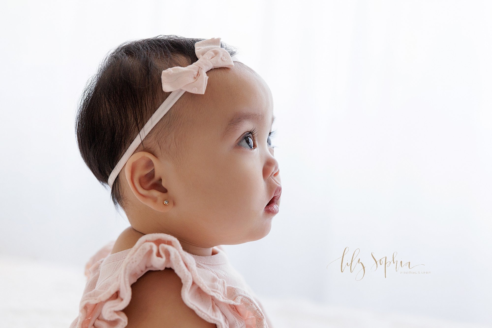  Profile portrait of a one year old Asian little girl for her first birthday as she sits on a bed wearing a bow headband in her hair in a studio in front of a window streaming natural light near Old Fourth Ward in Atlanta, Georgia. 