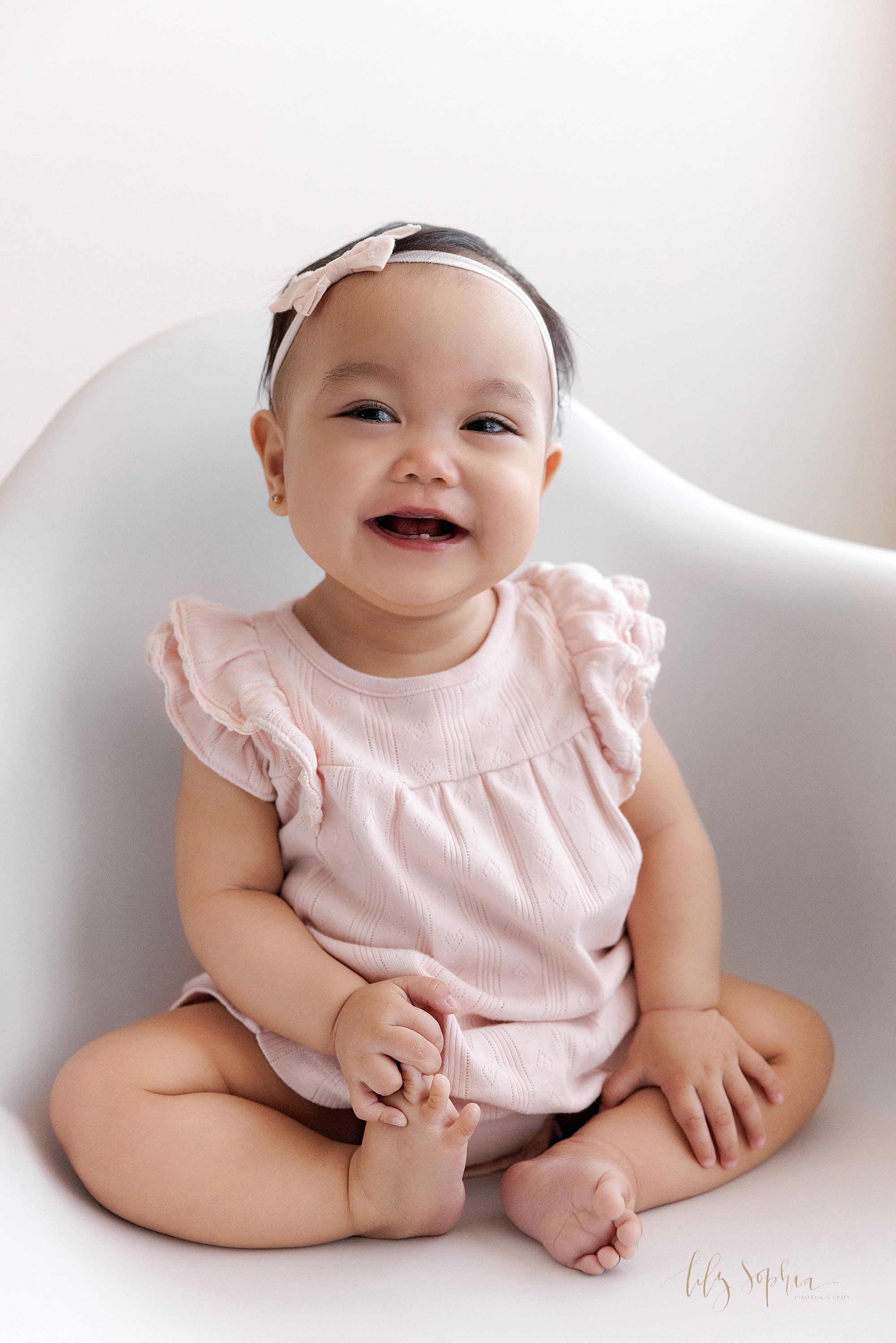 First birthday photo session of an Asian one year old girl as she sits in a white molded chair smiling to show her tiny baby tooth while playing with her right foot as natural light streams in from a window in a photography studio near Midtown in At