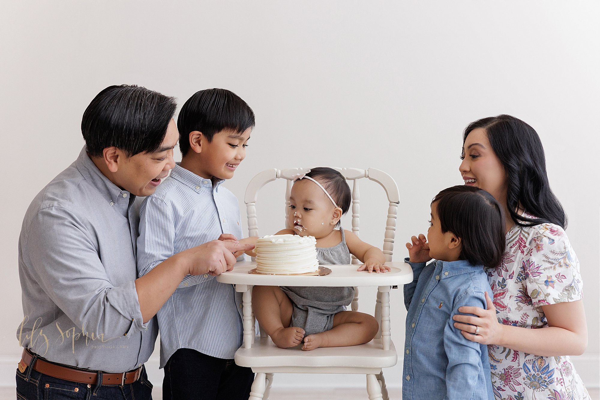 Family first birthday portrait of a one year old Asian girl surrounded by her brothers and parents as she sits in an antique high chair with icing on her face from the smash cake sitting on the tray taken near Roswell in Atlanta in a photography stu